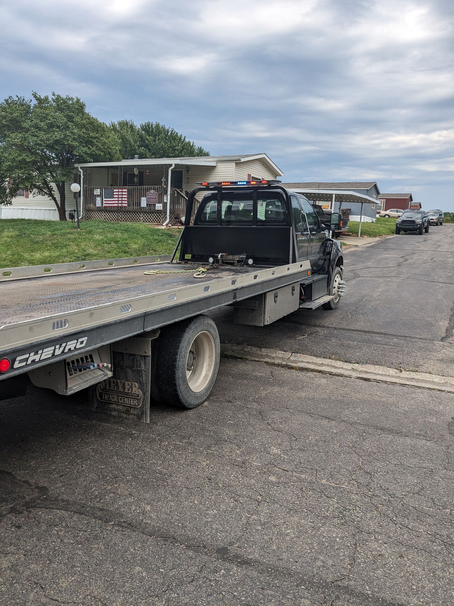 A black flatbed tow truck parked on a paved road beside mobile homes under a cloudy sky.