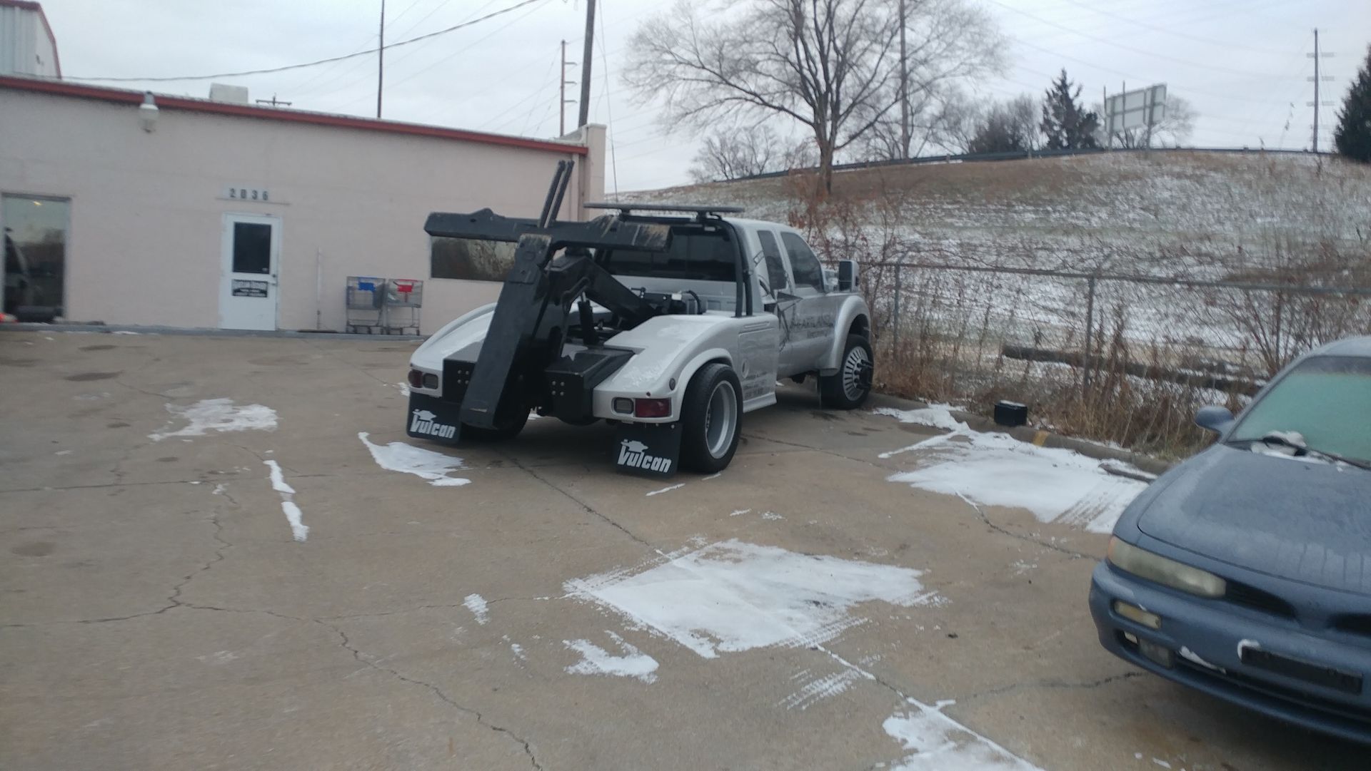 A silver tow truck parked in a snowy lot next to a light-colored building and a parked sedan.