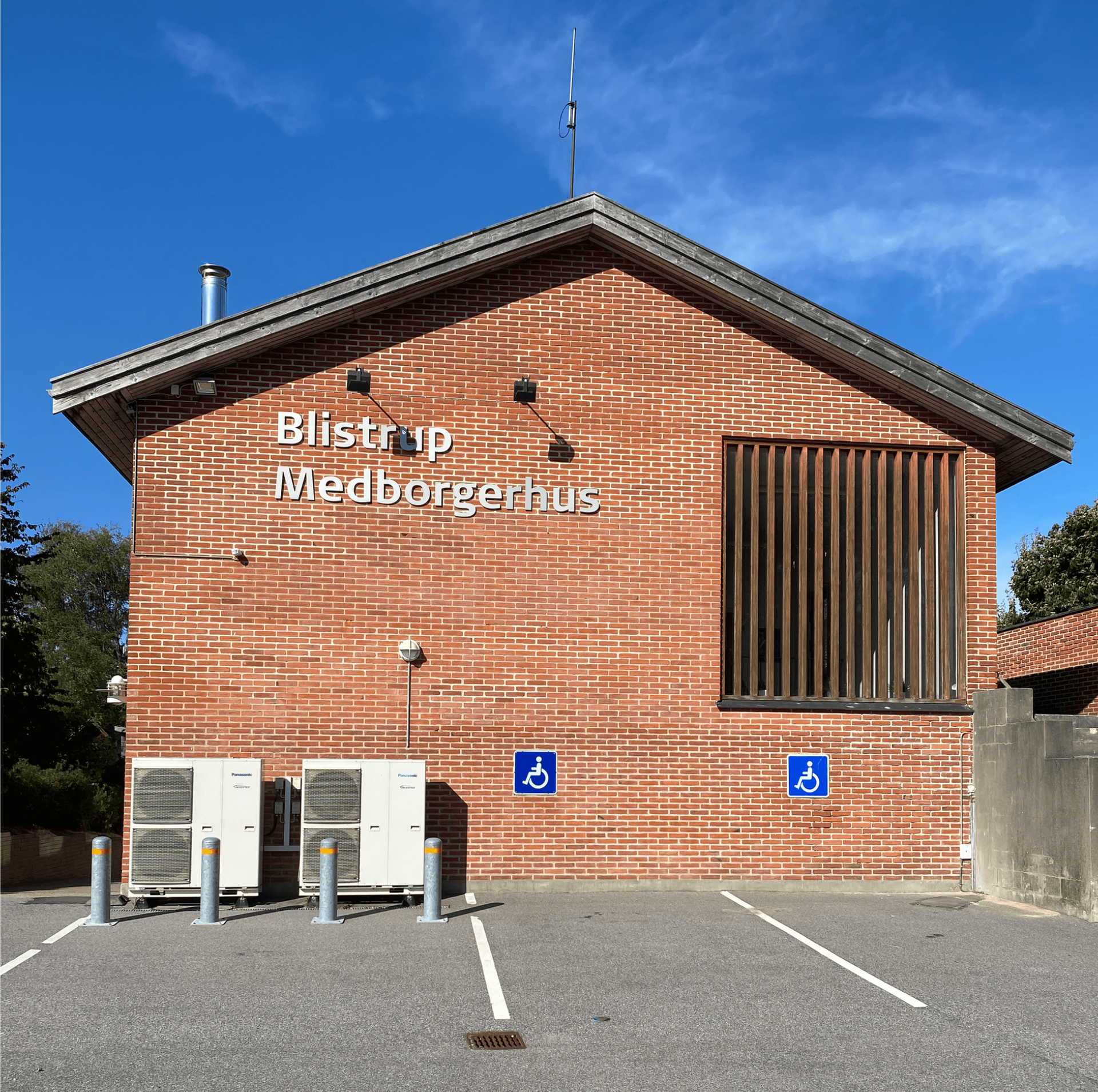 Blistrup Medborgerhus, murstensbygning med blå handicapskilte og parkeringspladser på en solskinsdag.