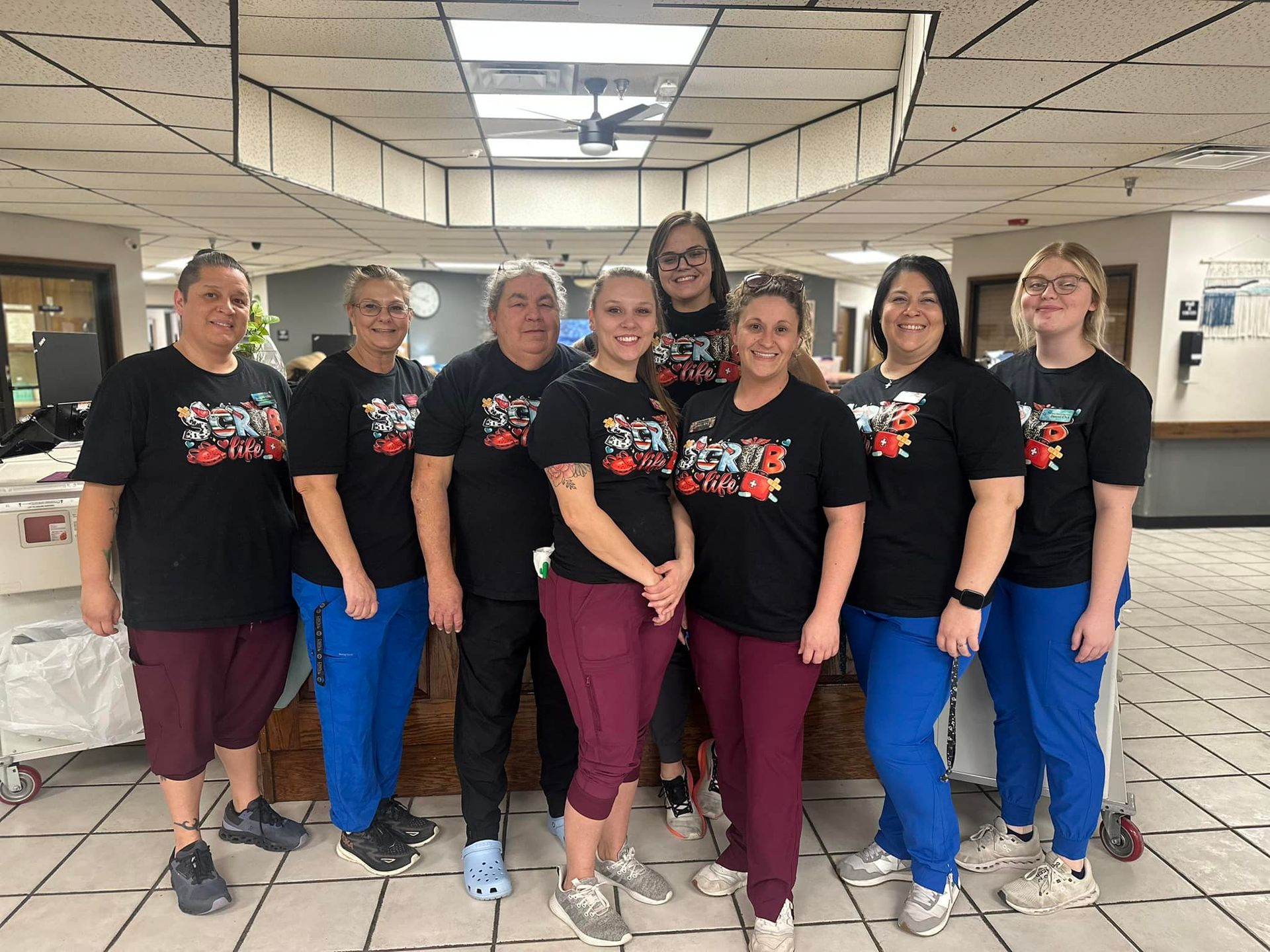 Group of eight healthcare workers in black t-shirts and scrubs smiling, posing indoors.