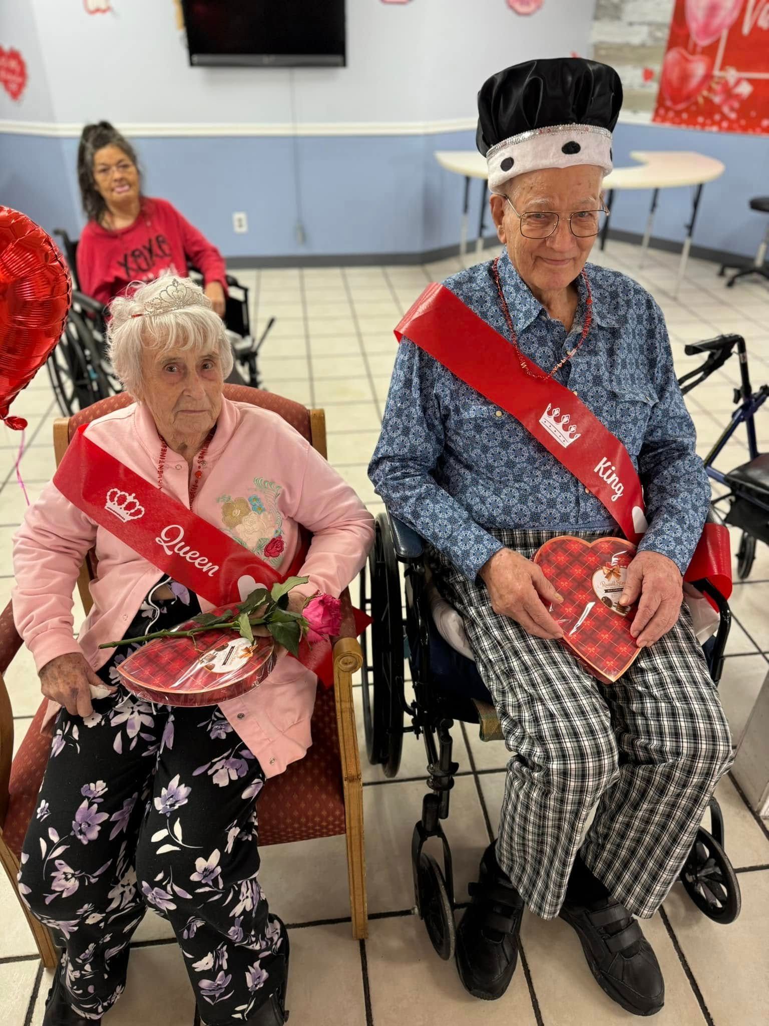 Senior couple, Queen and King, with sashes and crowns, holding heart treats. A woman smiles behind them.