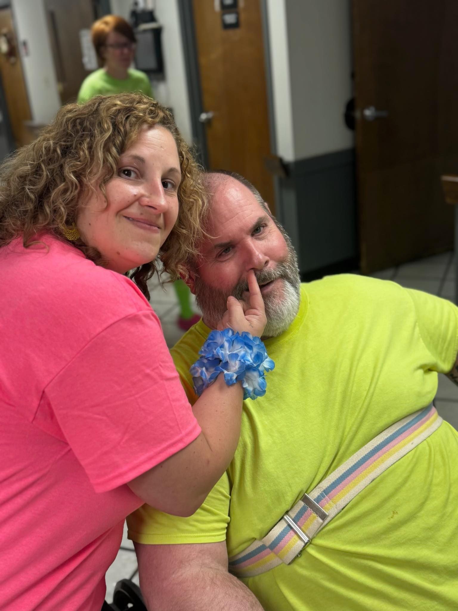 Woman in pink shirt touches man's face with blue flower. Man in yellow shirt sits, smiling. Third person in green shirt in background.