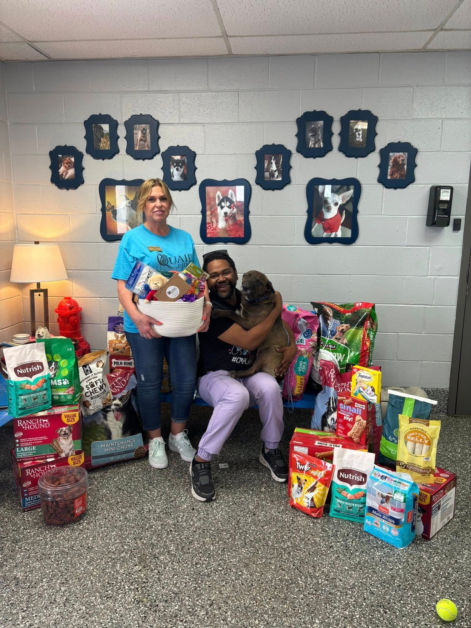 Two people with dog and donations for animals. Indoor setting, beige walls, food bags.