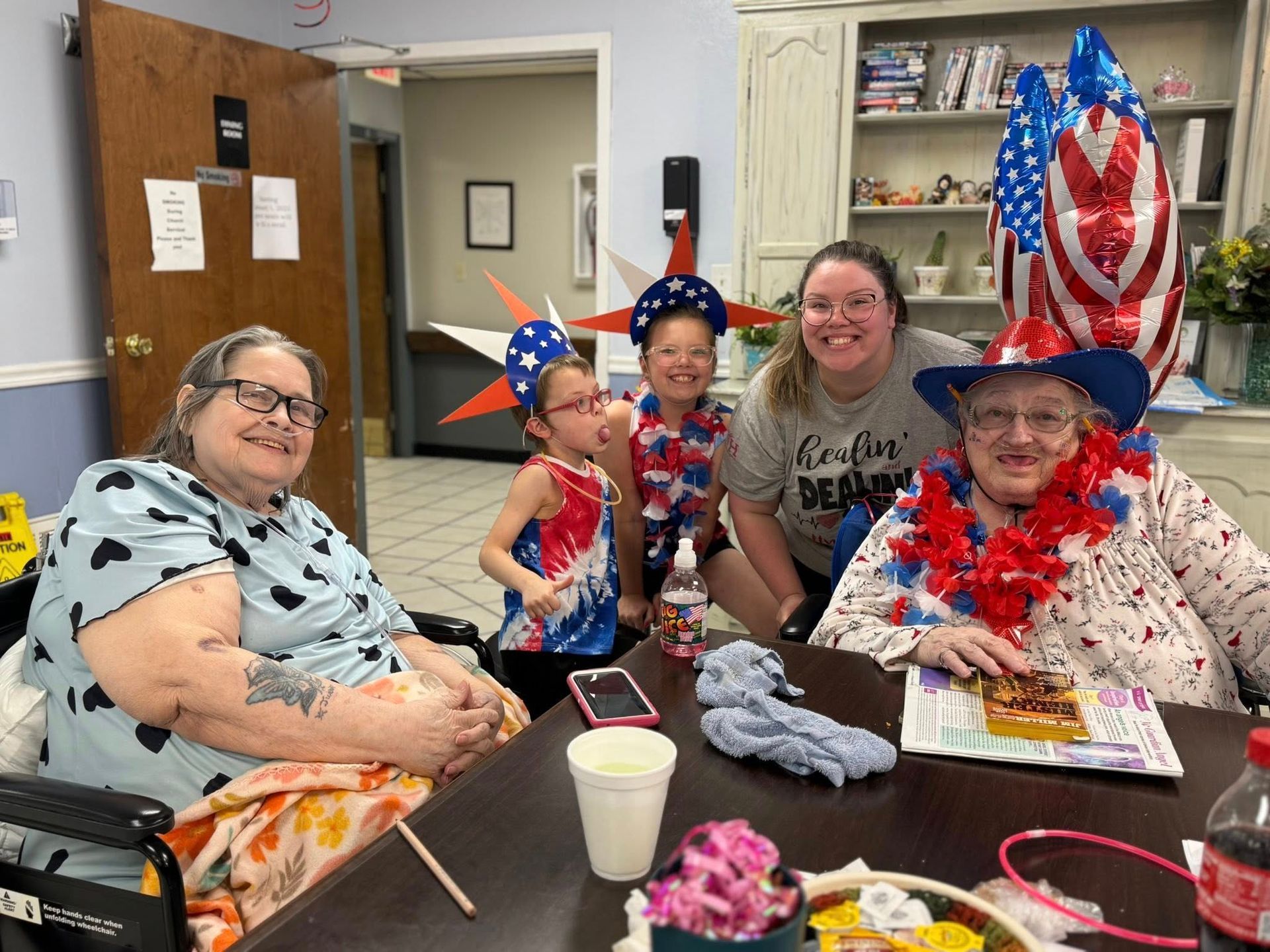Group celebrates with patriotic decor. Two women in wheelchairs, two children, and an adult pose smiling at a table.