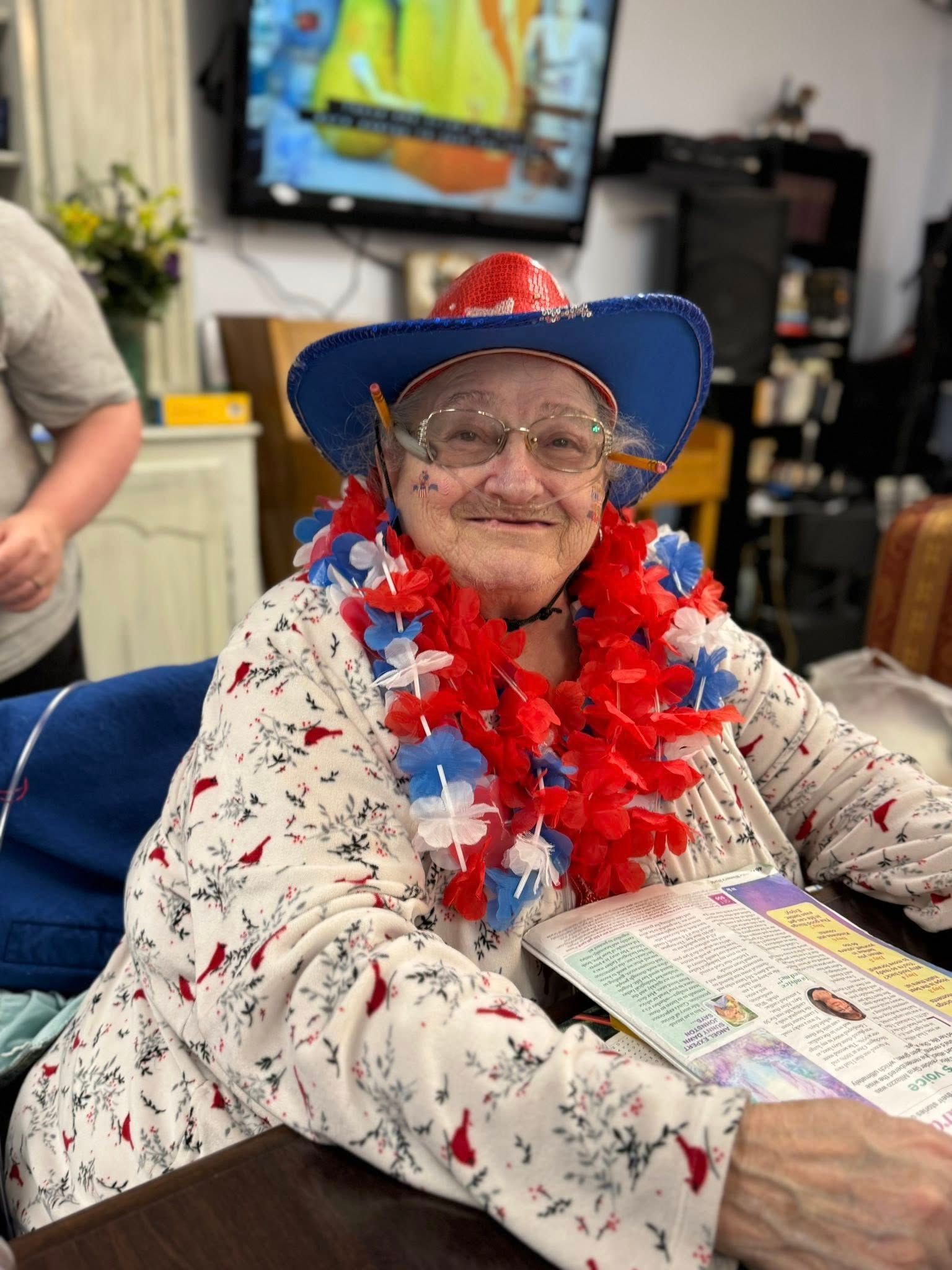 Elderly woman wearing a patriotic hat and lei, smiling while holding a paper in a room.