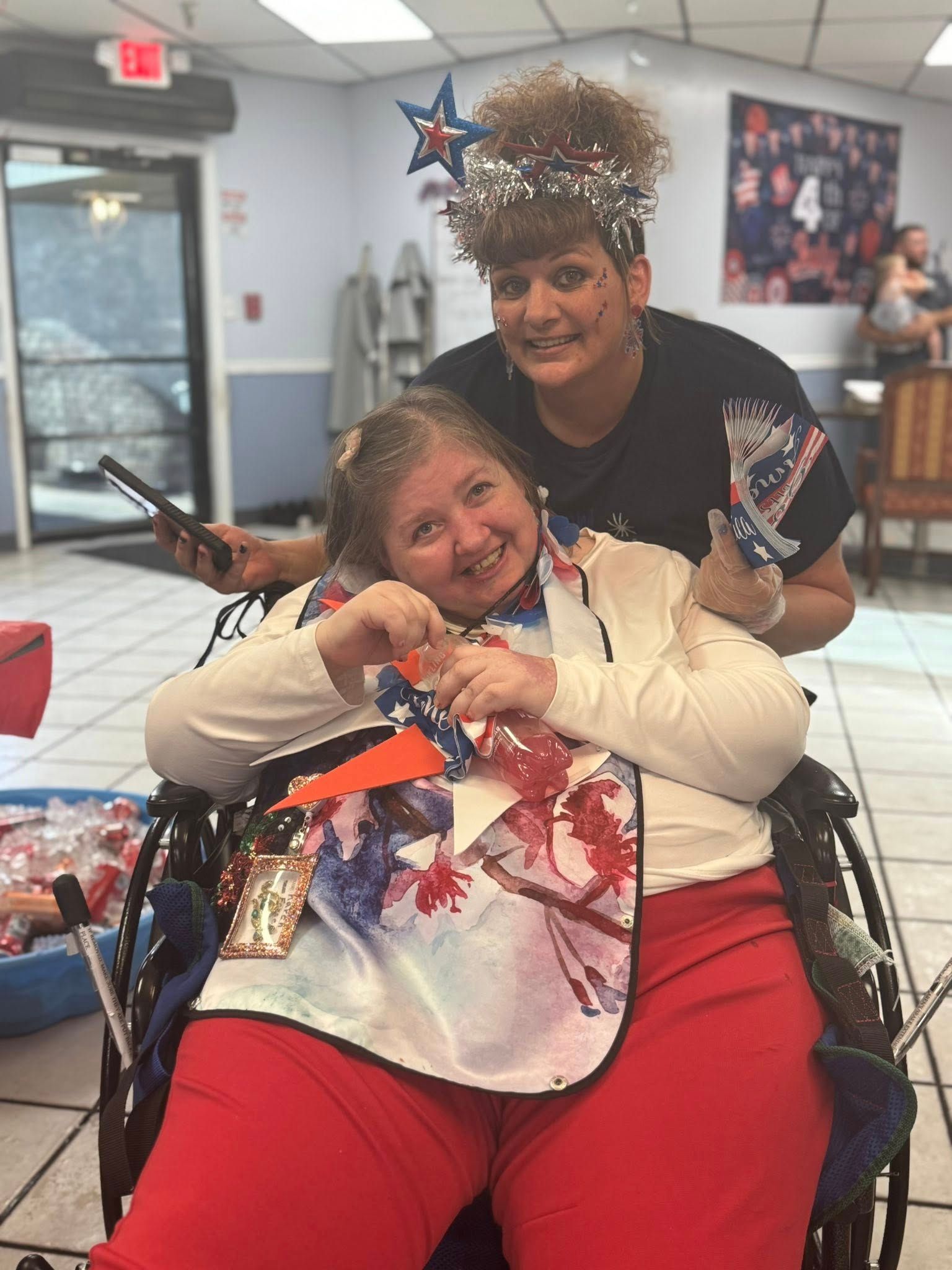 Two women smiling; one in wheelchair, wearing patriotic attire, other behind her, wearing a star headband, indoors.