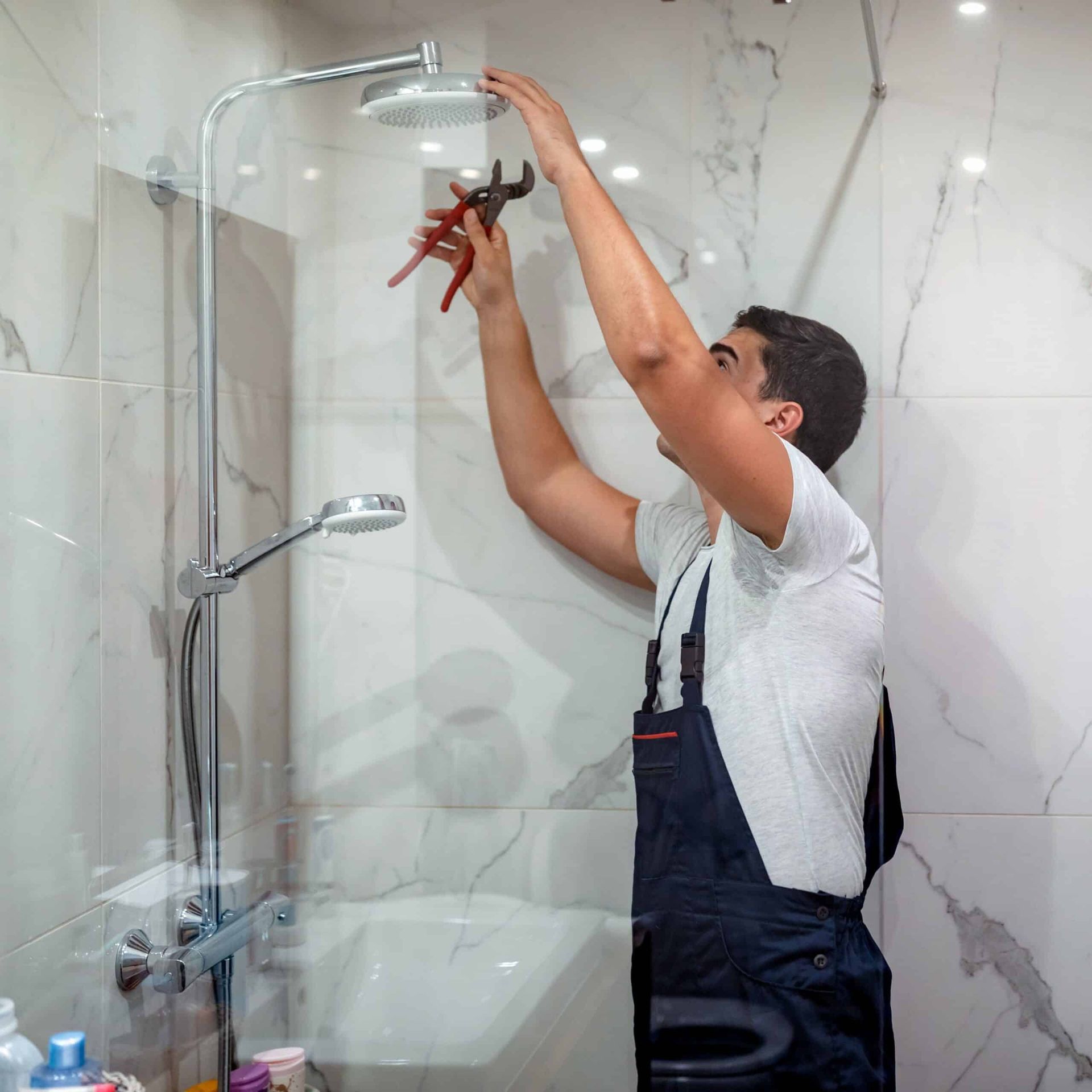 Un plombier en salopette bleue utilise une pince pour réparer un pommeau de douche dans une salle de bains carrelée de blanc.