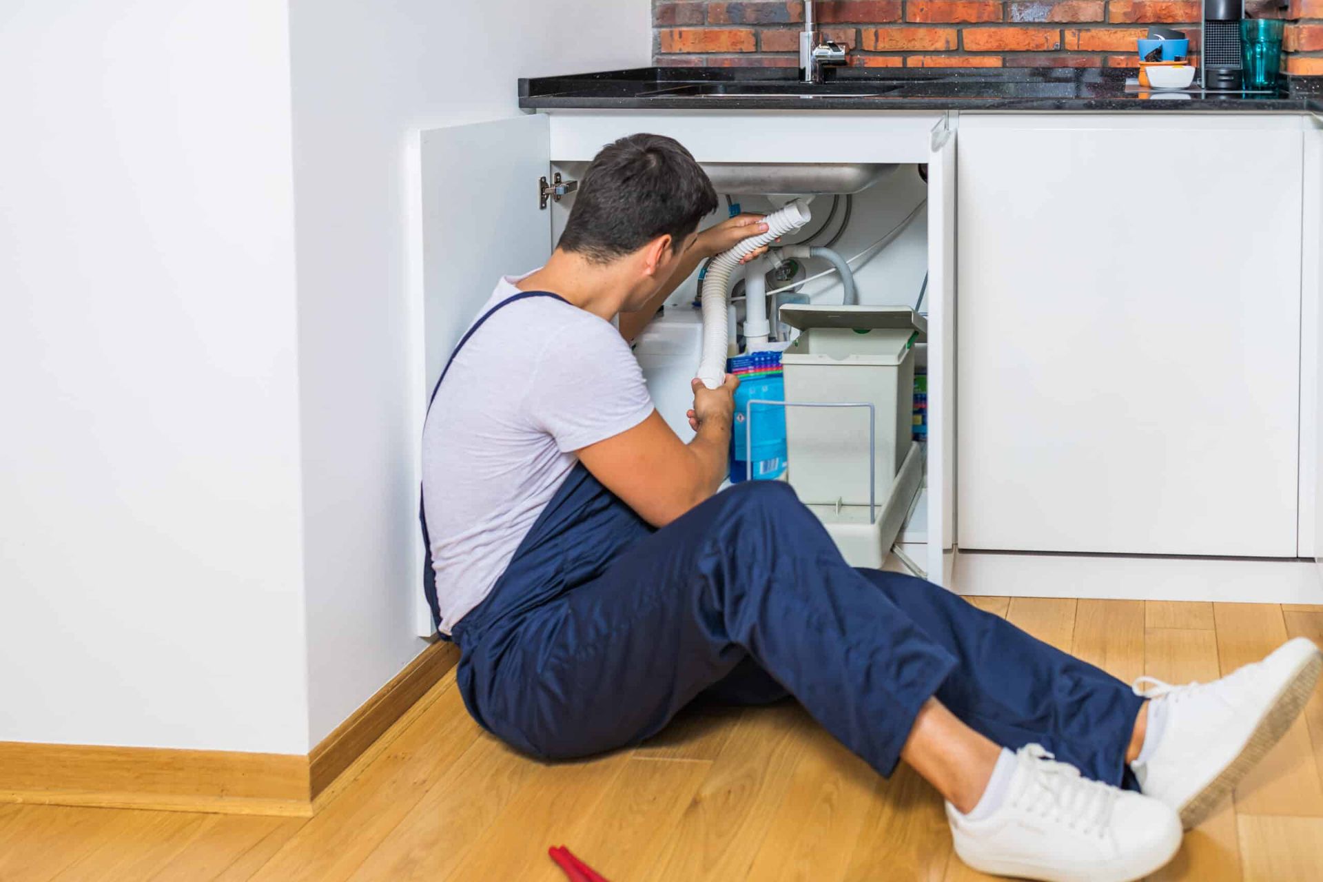 Une personne en salopette travaille sous un évier, en train de réparer la plomberie. Elle porte une chemise blanche et tient un outil. Scène de cuisine.
