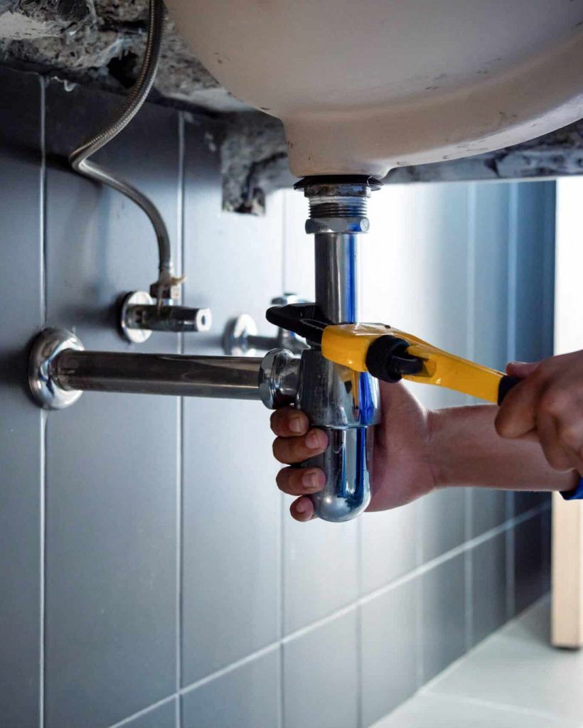 Un plombier utilise une clé sur un tuyau chromé sous un évier, dans une salle de bains carrelée de gris.