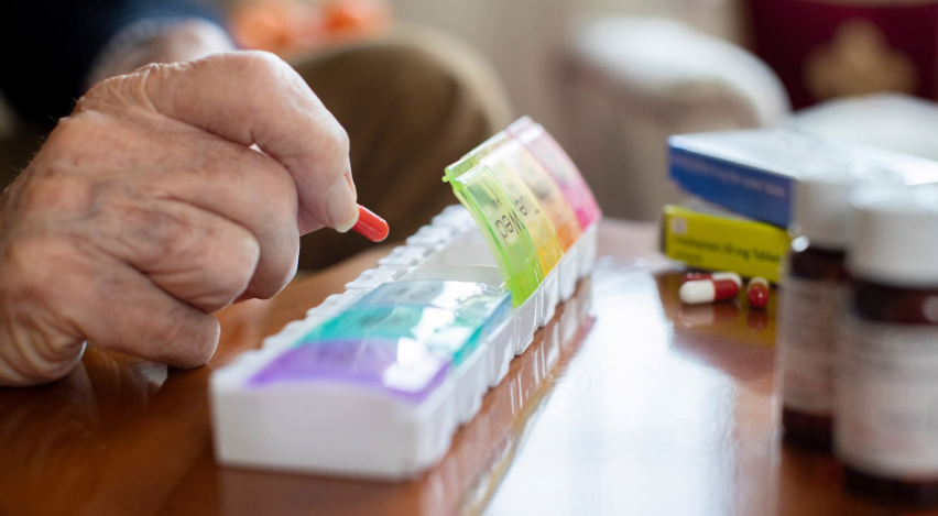 A hand places a red pill into a weekly medication box, surrounded by pill bottles.