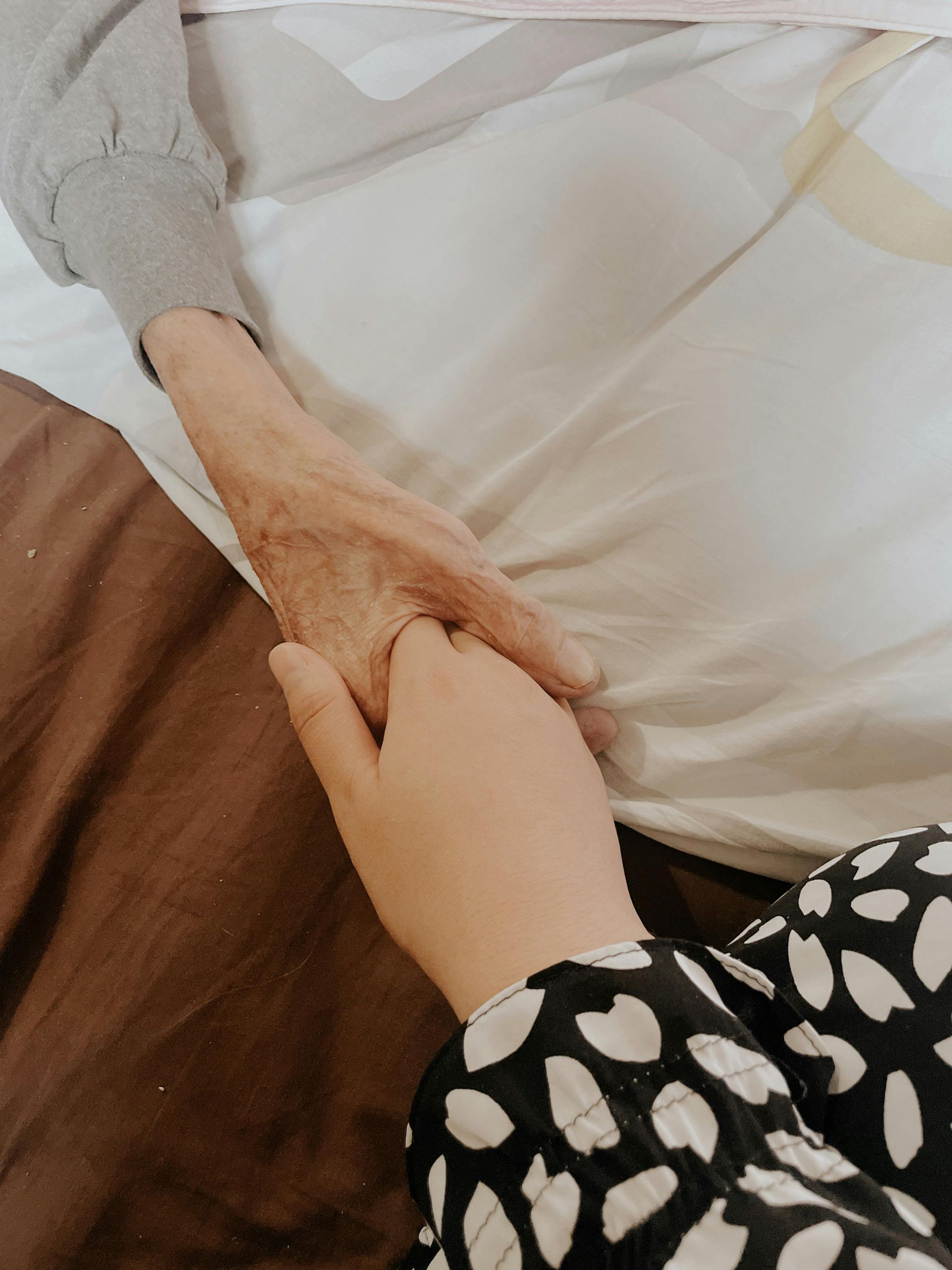 An older hand grasps a younger hand above a brown bed blanket