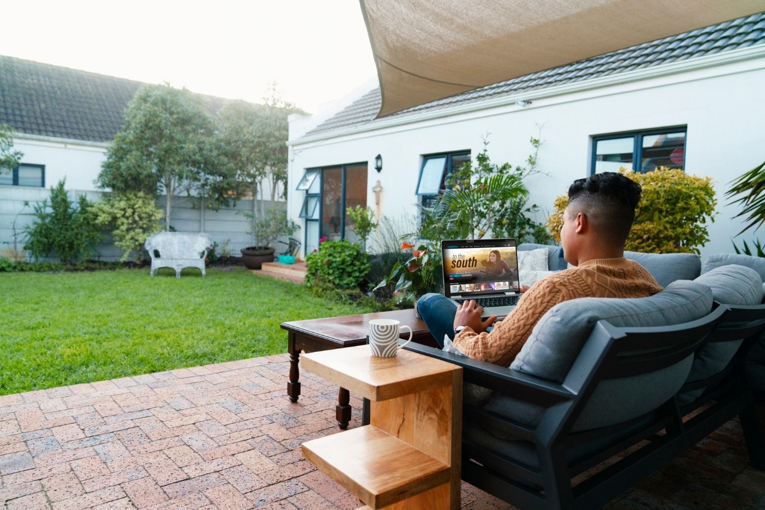Man sitting on patio couch using a laptop, outdoors in backyard.