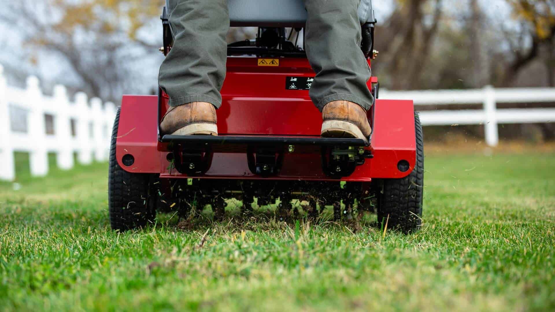 Person riding a red lawn aerator on green grass near a white fence.