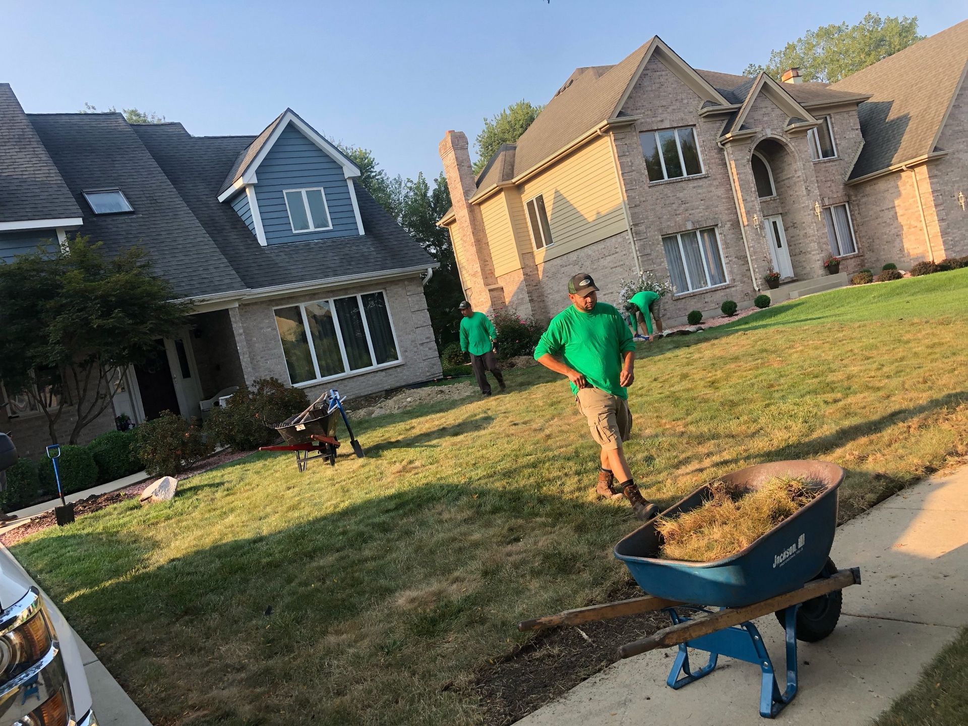 Lawn care workers mulching a front yard in front of two-story houses. One worker stands with wheelbarrow.