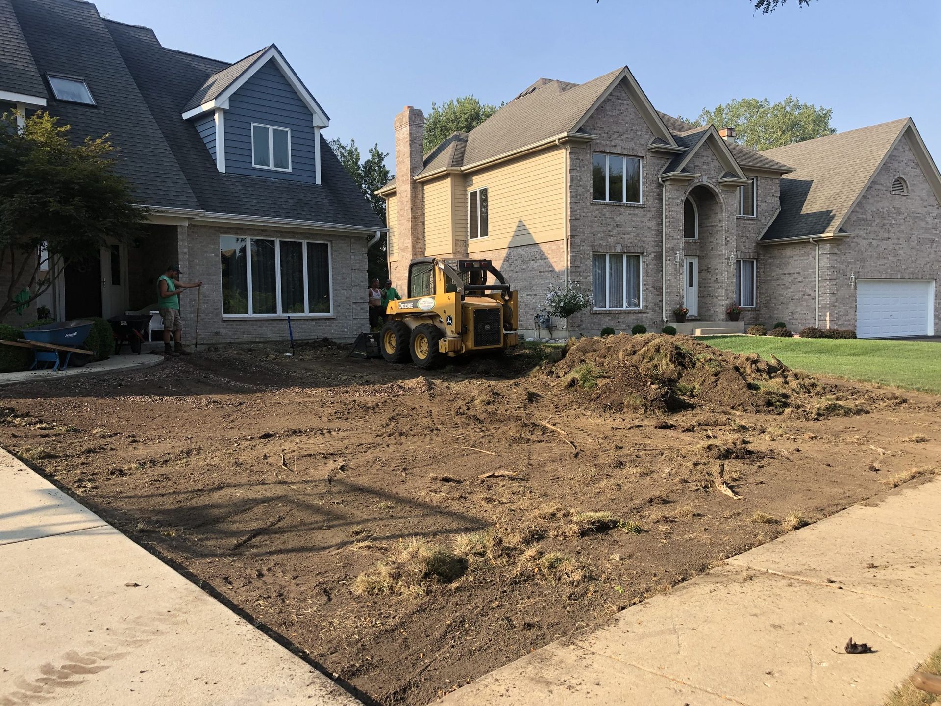 Skid steer on a dirt lawn between two houses, preparing ground for landscaping.