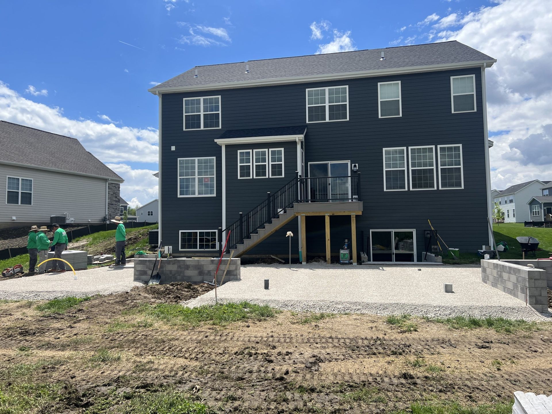 Rear view of a two-story blue house with deck overlooking a gravel patio under construction; workers nearby.