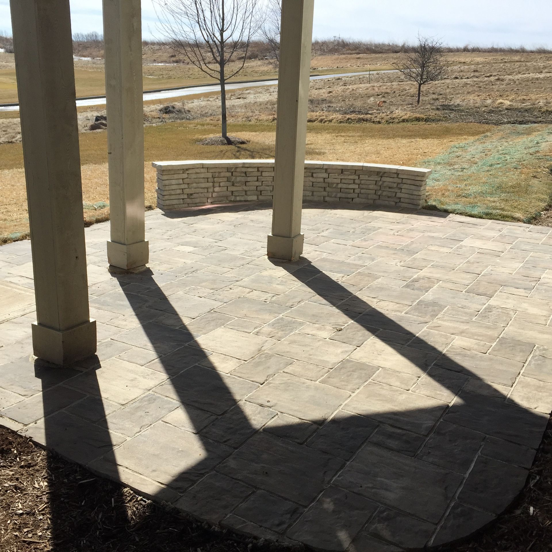 Stone patio with three pillars casting shadows, a low stone wall, and a grassy field in the background.