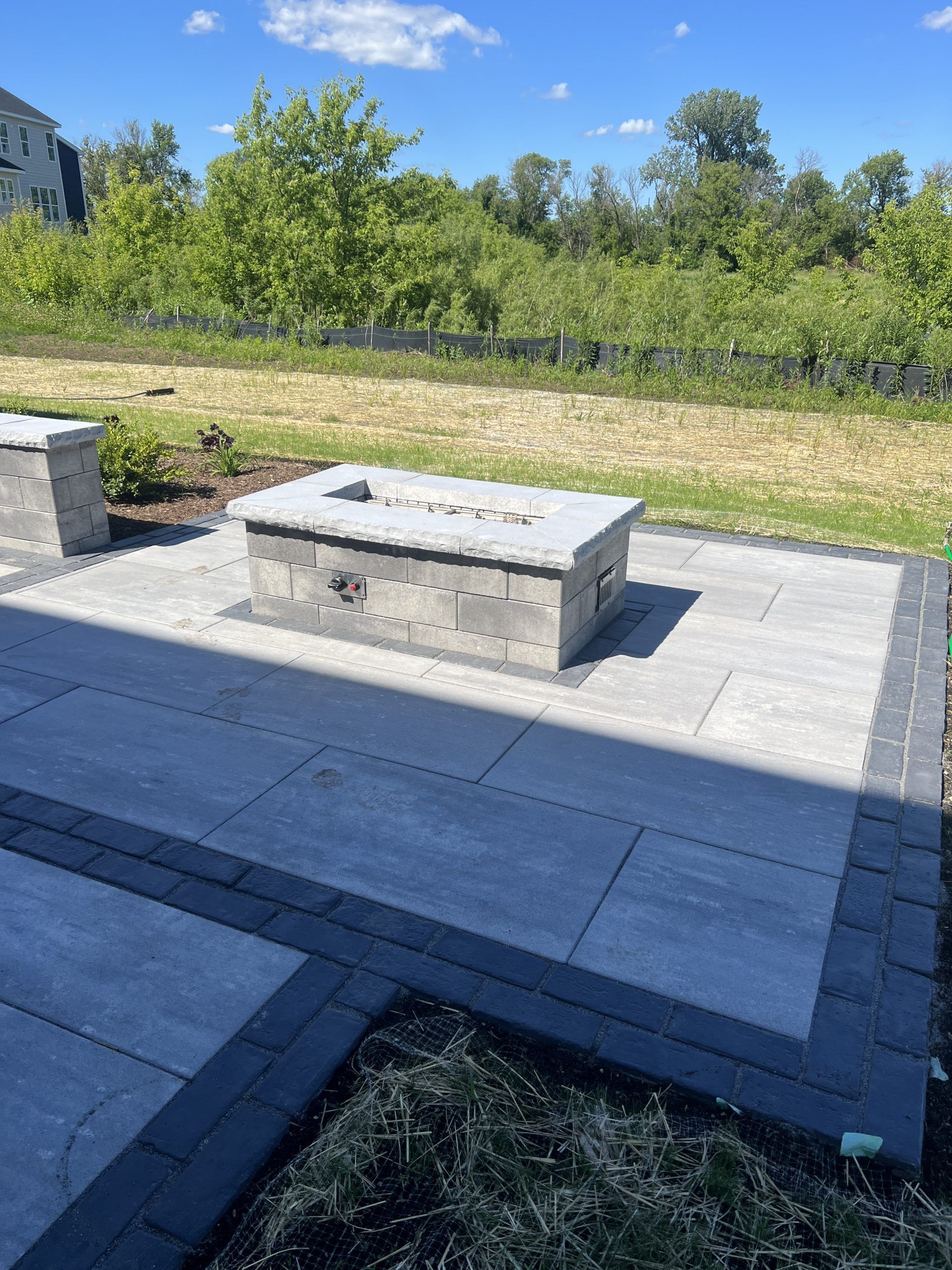 Stone patio with a fire pit, surrounding grass and trees under a sunny blue sky.