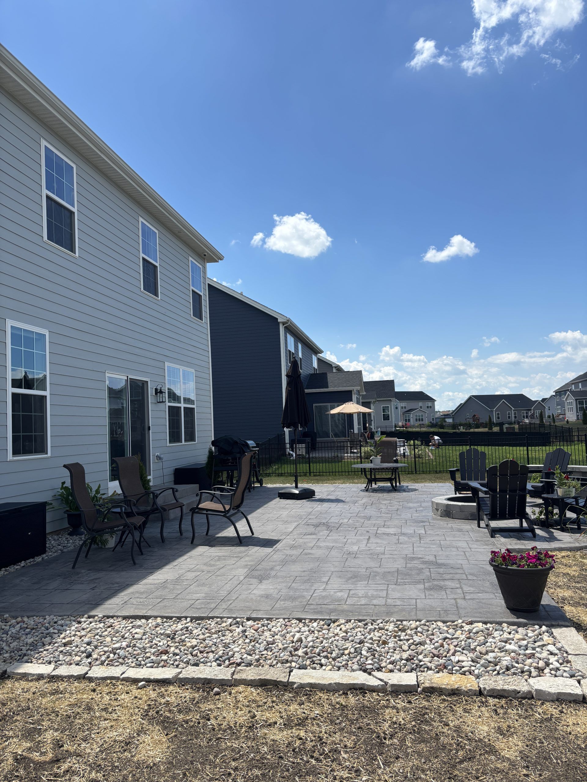Backyard patio with gray pavers, various chairs, and a small umbrella on a sunny day.