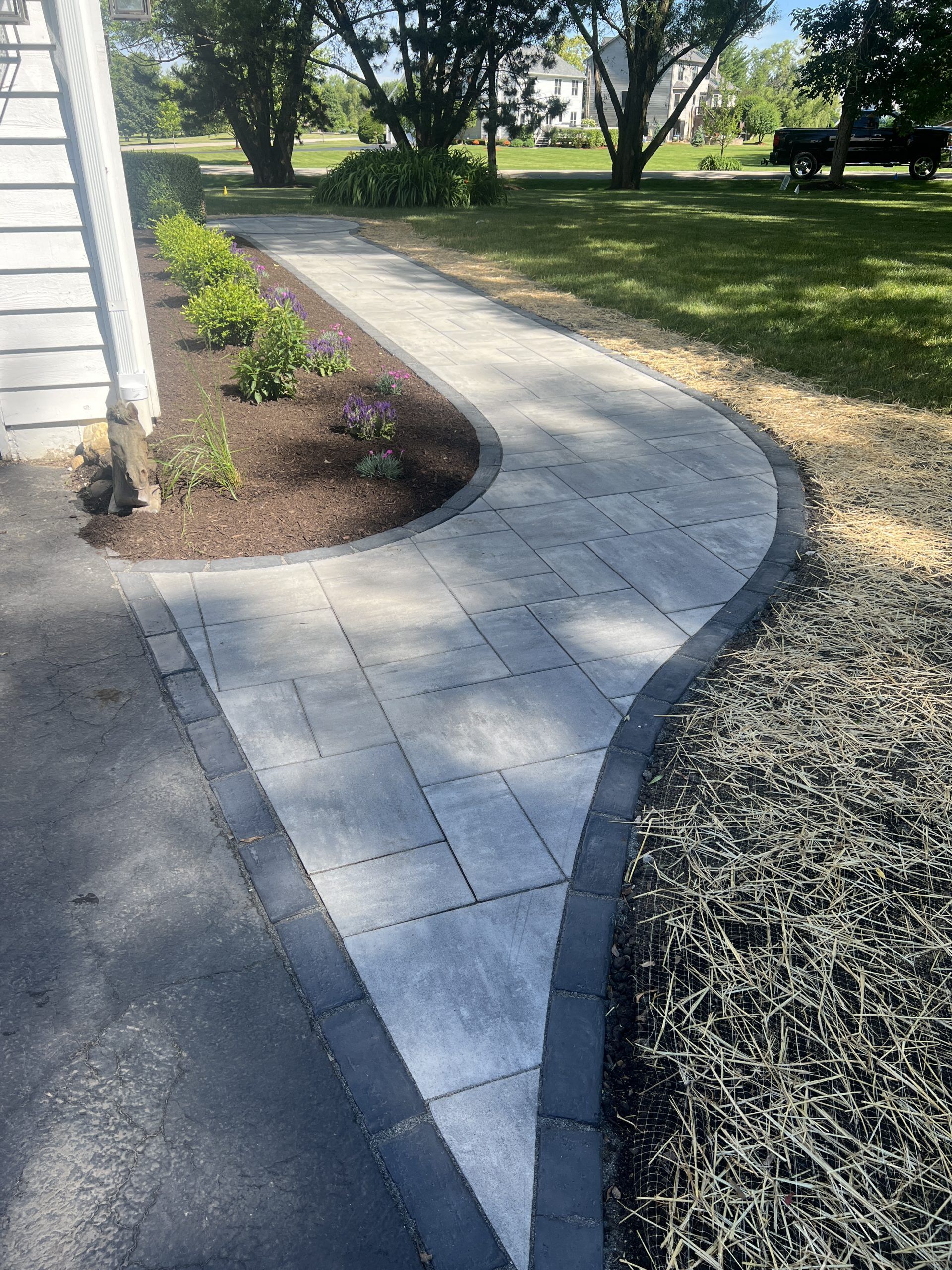Stone pathway curves through a yard, bordered by dark pavers and landscaping near a house.