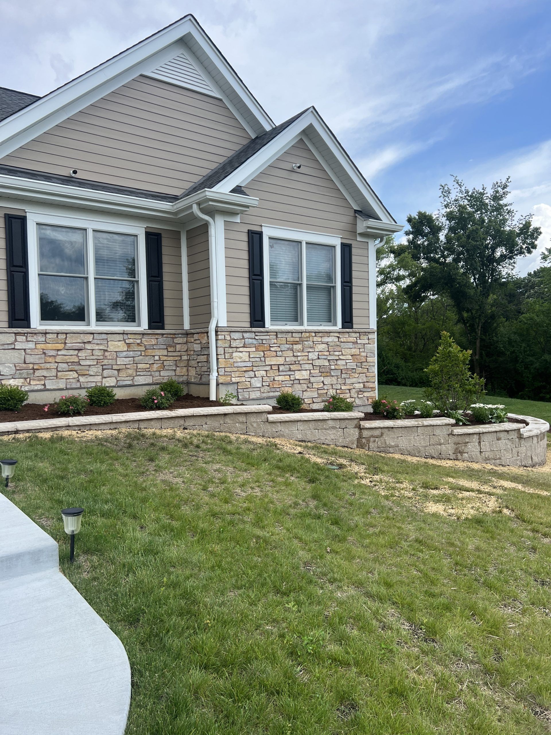 Beige and stone veneer house with black shutters, surrounded by grass and a low retaining wall.