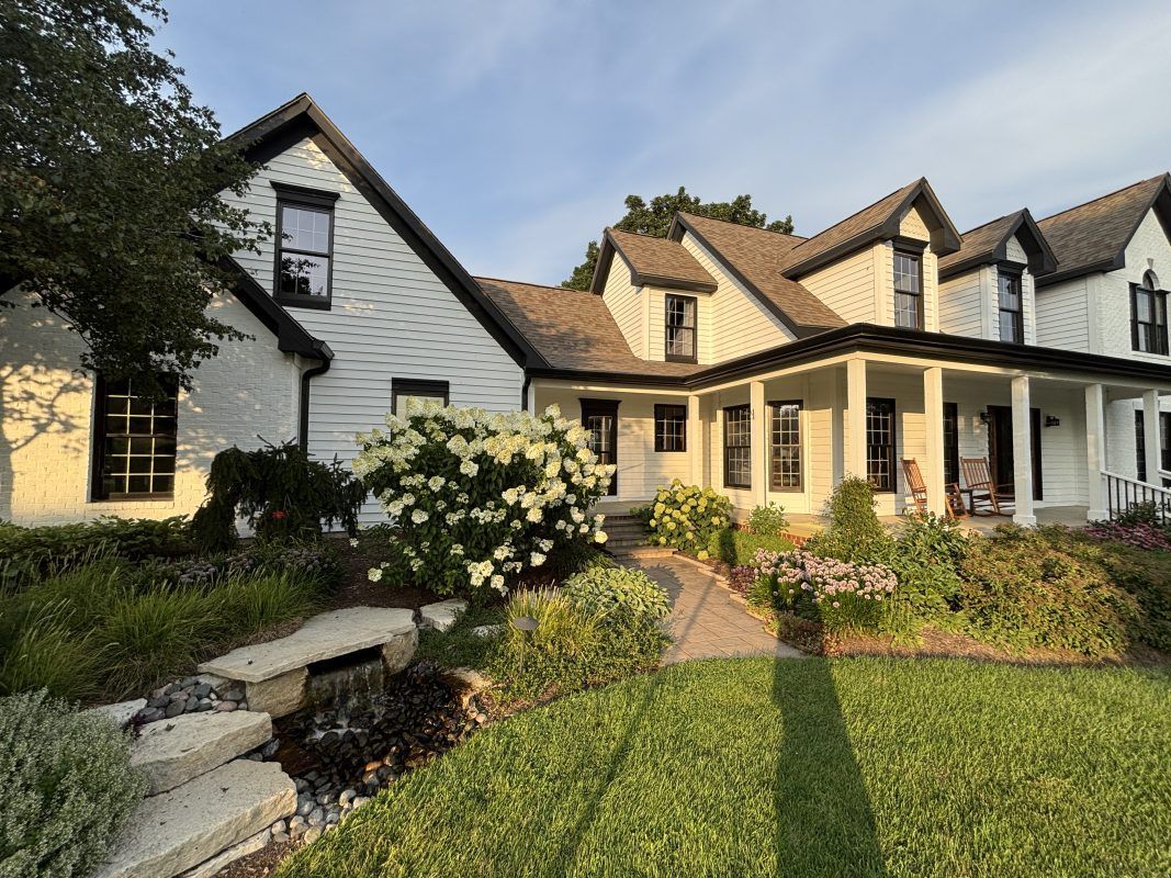 White farmhouse with black trim, a long porch, and a lush garden.
