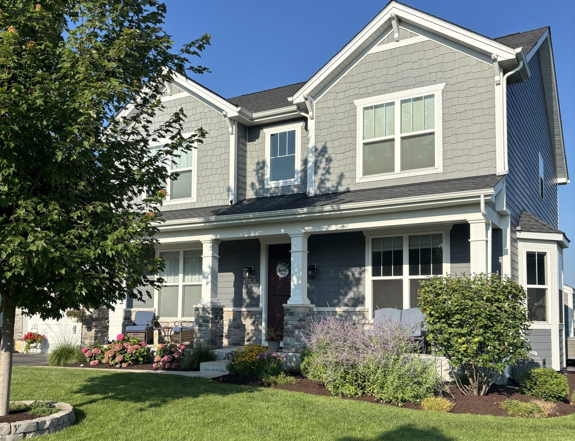 Gray two-story house with white trim, porch, and landscaping under a clear blue sky.