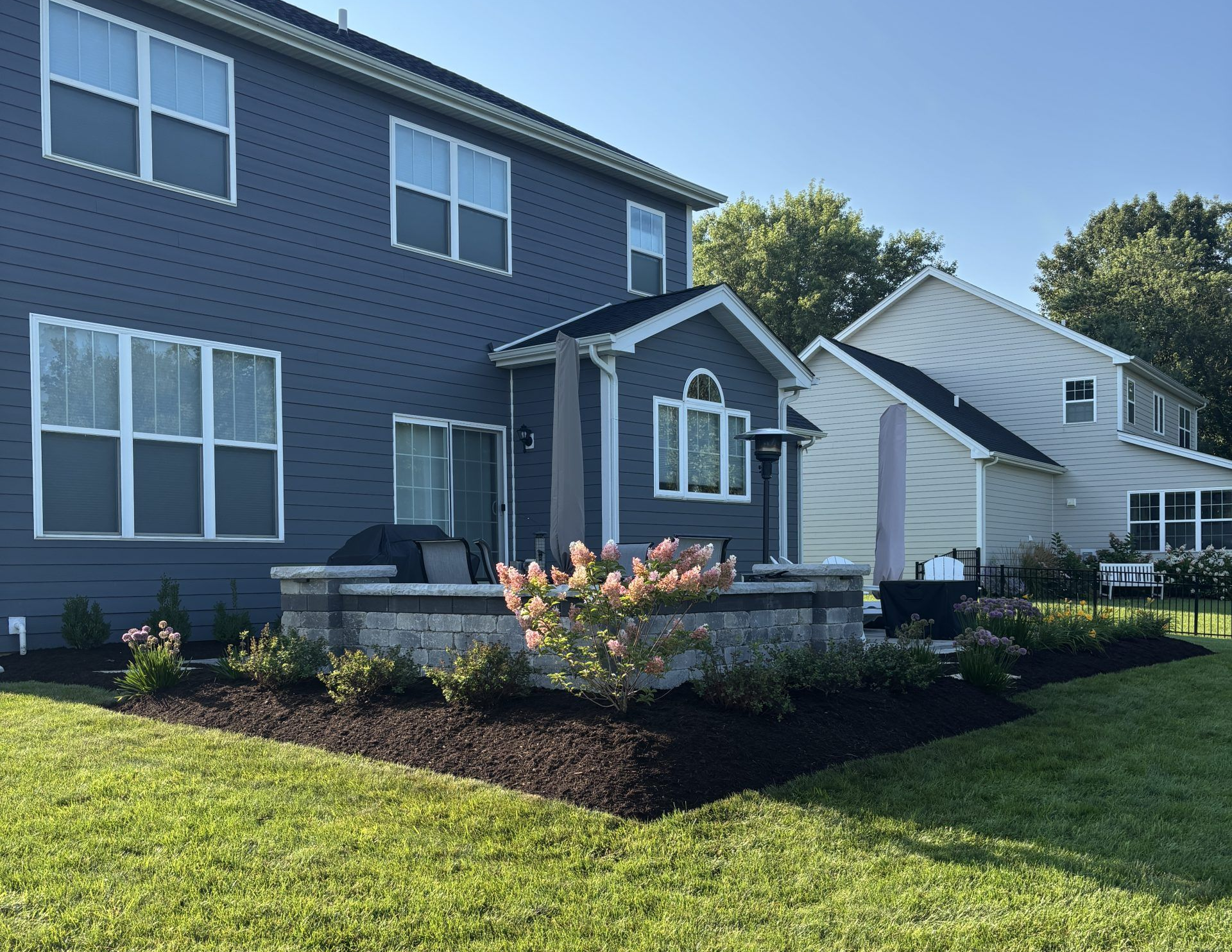 Backyard with blue house, stone patio, green lawn, and flower beds under a sunny sky.