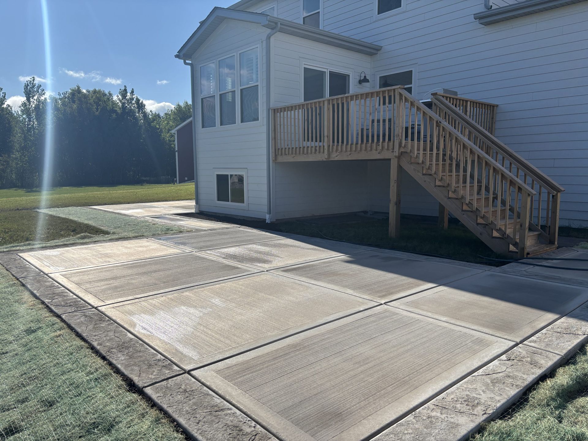Backyard patio with concrete slabs, wooden deck, stairs, and house. Green grass surrounds.