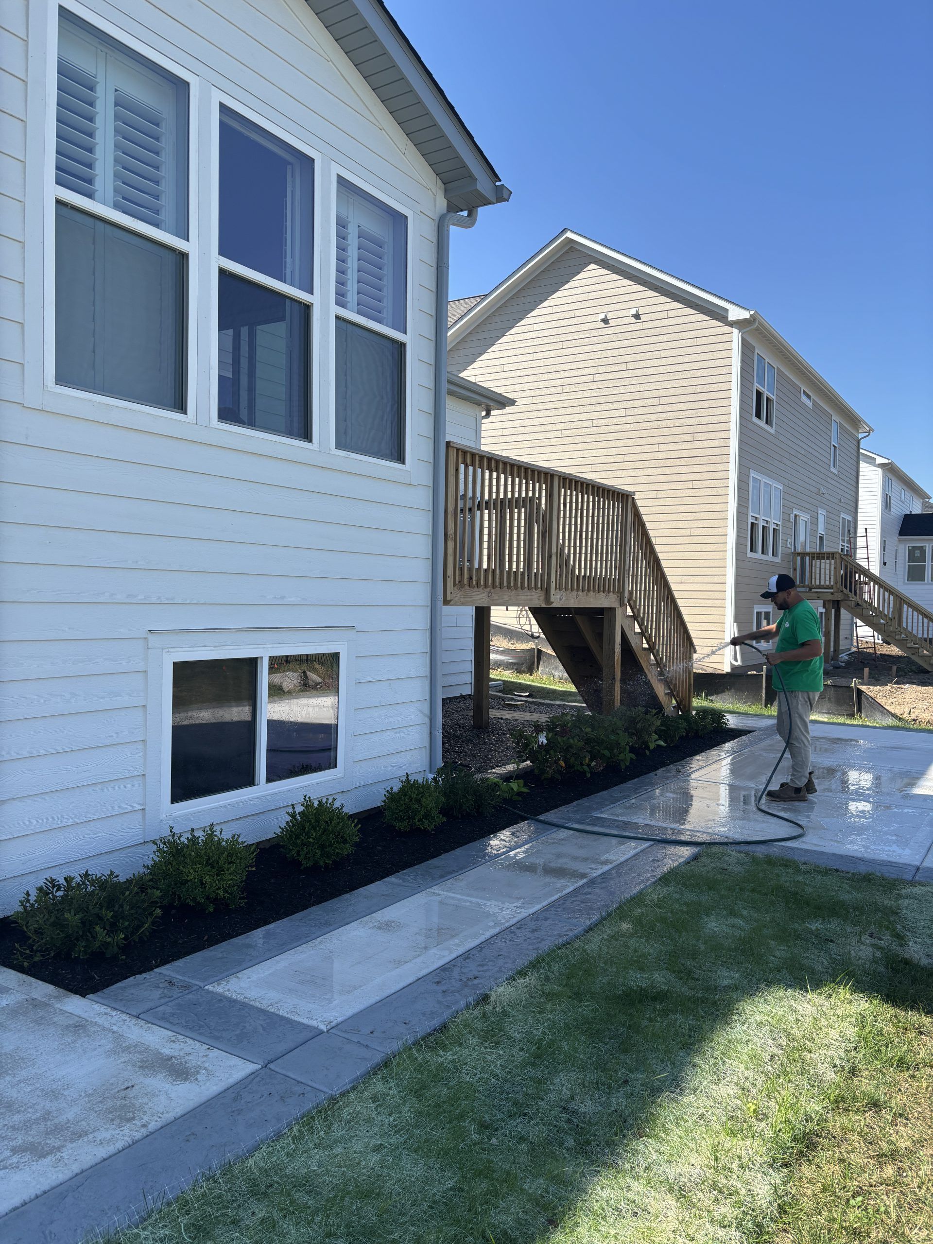 Man power washing a concrete patio next to a house with white siding and a wooden deck.