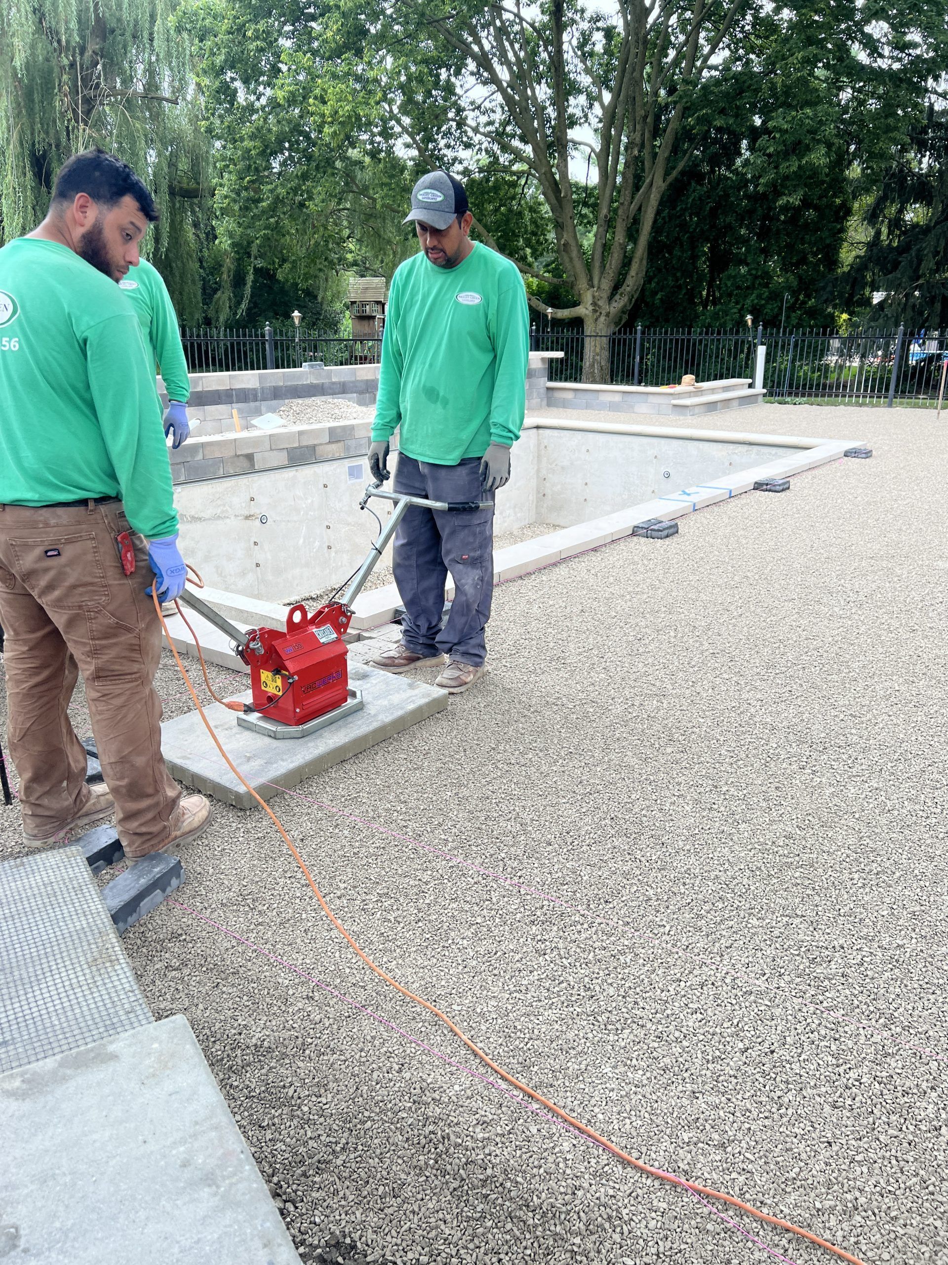 Three workers in green shirts compact gravel around a pool using a vibratory plate compactor.