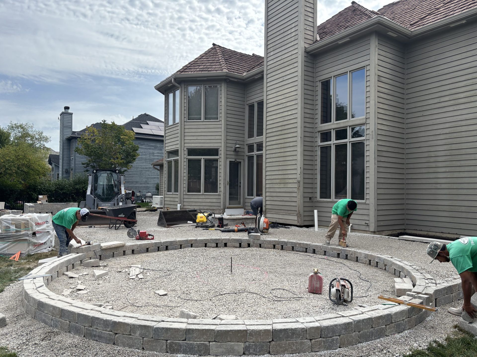 Workers constructing a circular patio with retaining walls near a large house.
