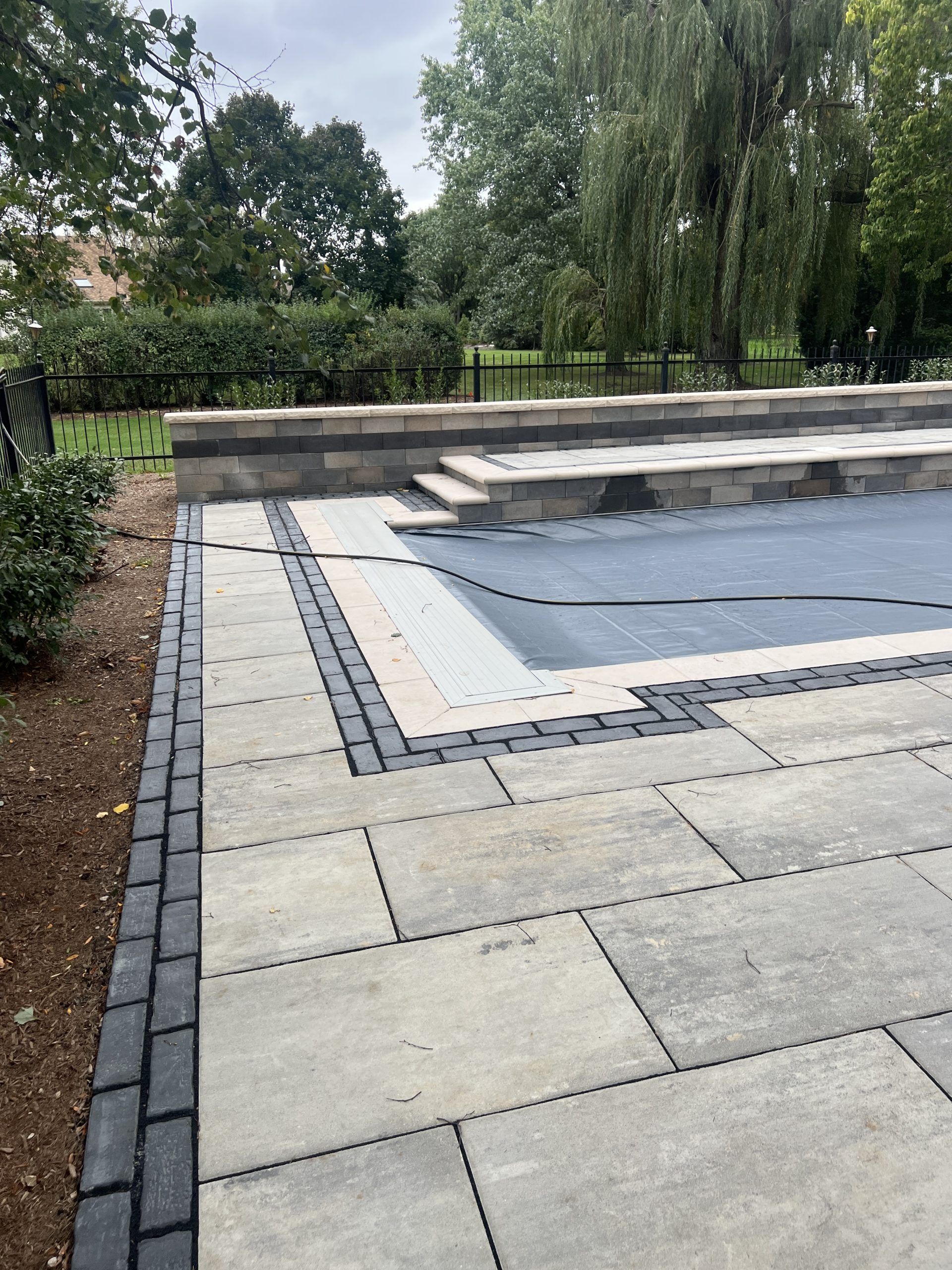 Patio with stone pavers and pool cover; dark border, low wall, steps, trees in background.