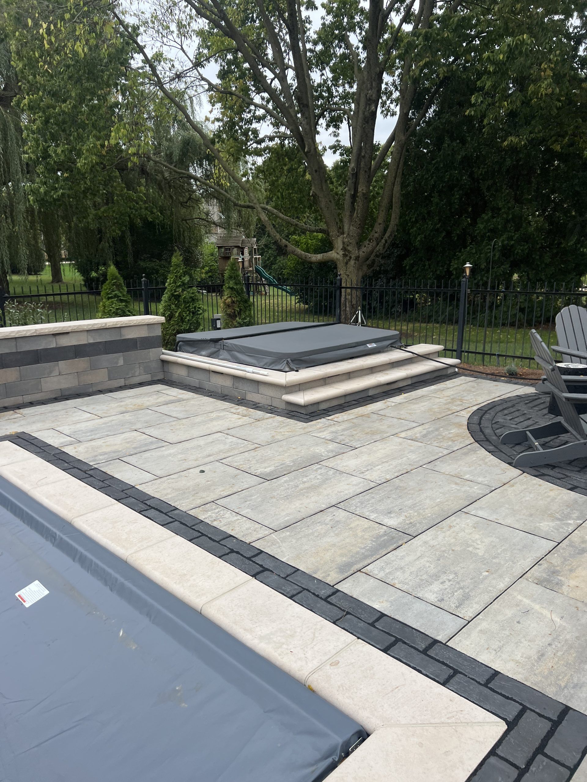 Stone patio with hot tub, steps, and pool in a yard with trees.