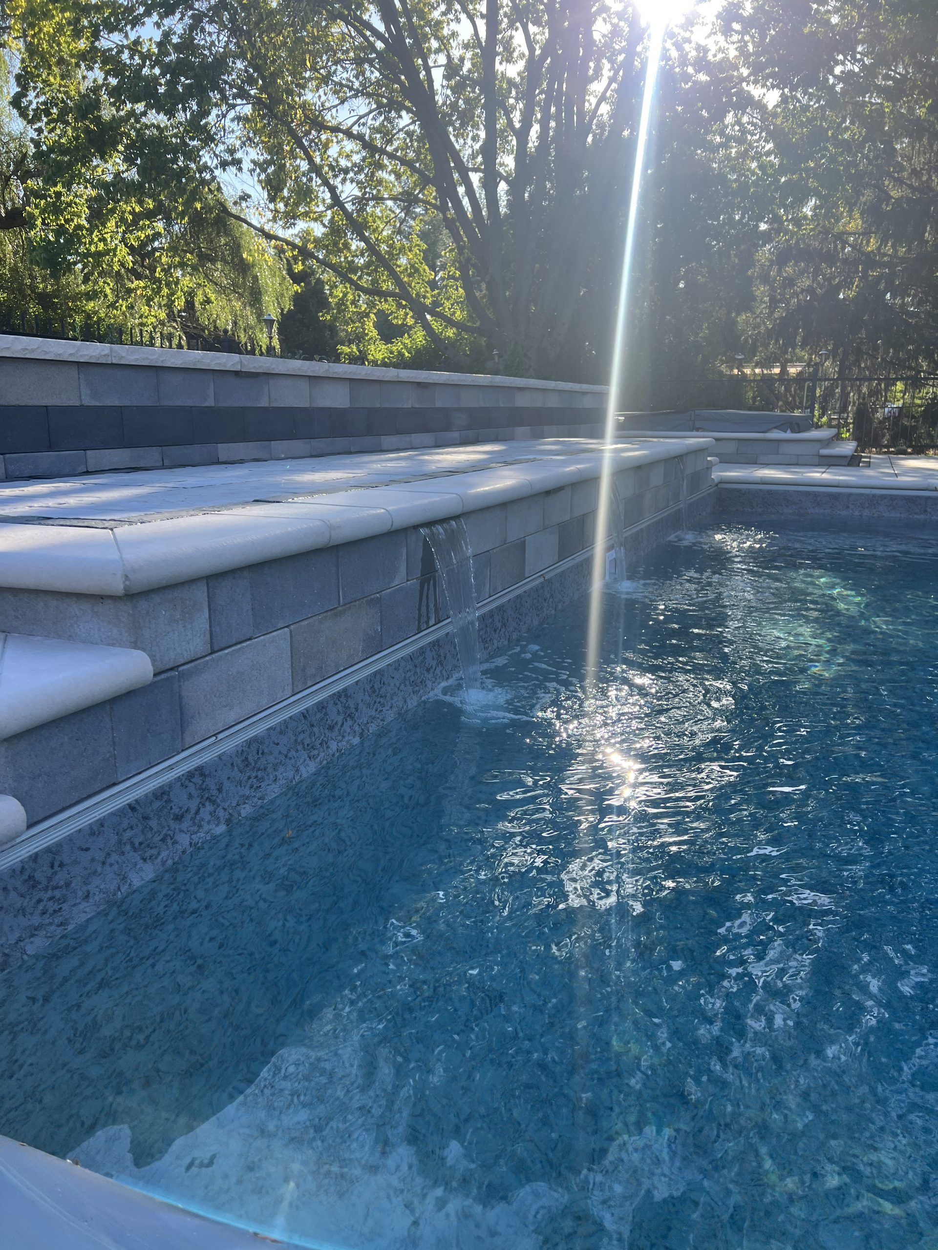 Swimming pool with blue water and sunlight reflecting, with a gray stone wall and water feature.