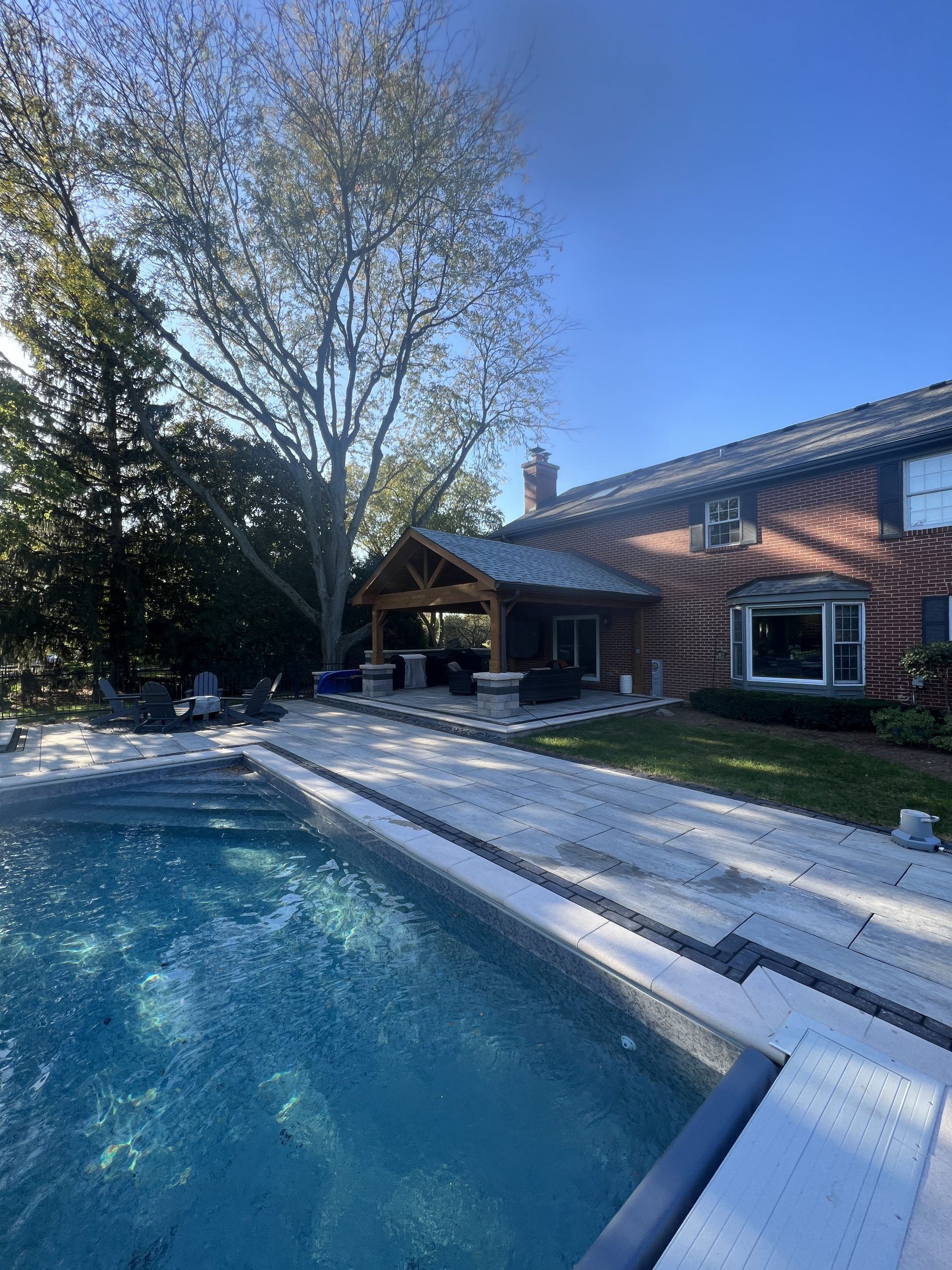 Backyard pool and patio with a brick house and large tree on a sunny day.