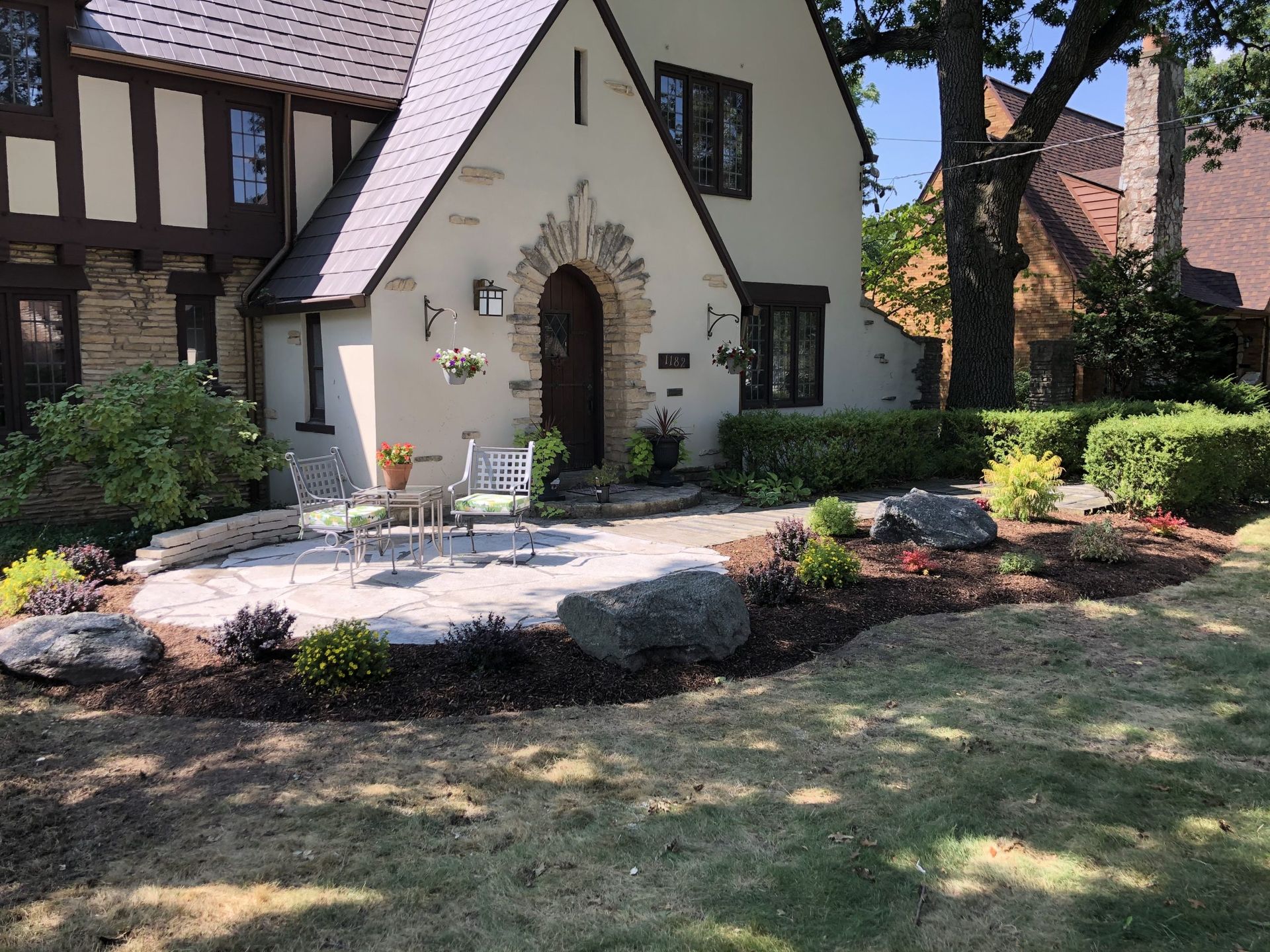 Stone patio in front of a Tudor-style house with manicured landscaping and large rocks.