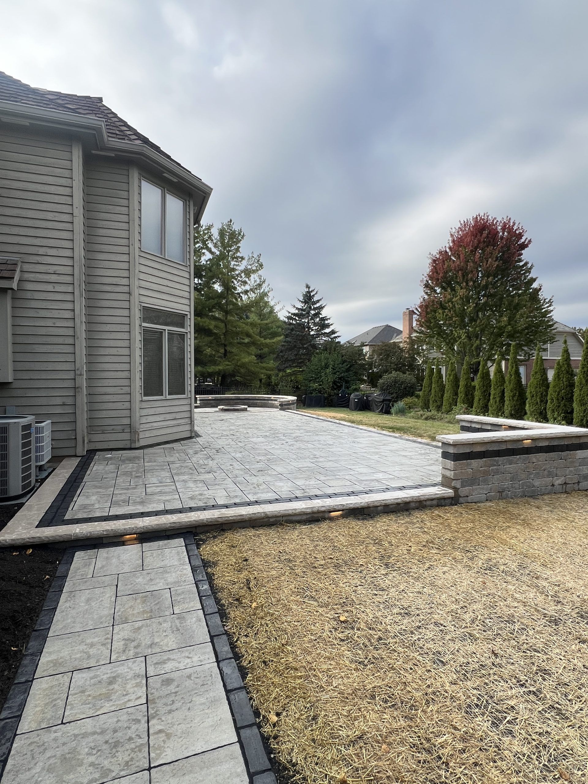 Backyard patio with gray pavers and a low retaining wall next to a house under a cloudy sky.