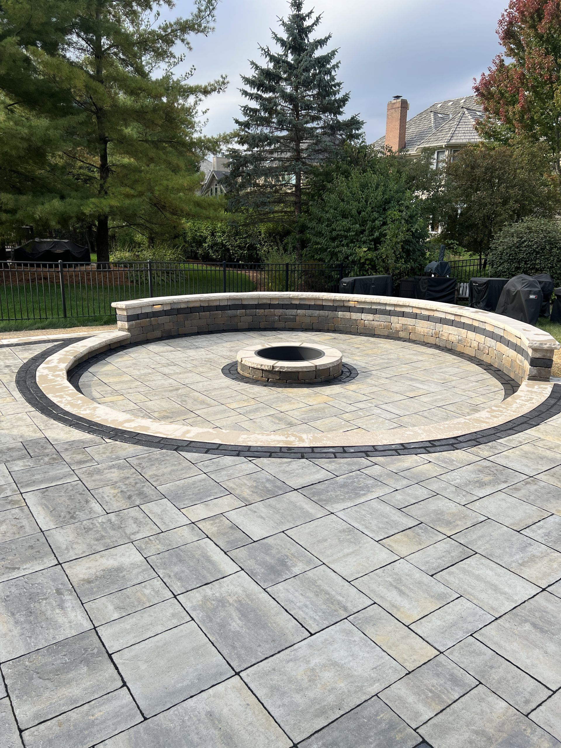 Circular stone fire pit in a paved patio, surrounded by trees and a cloudy sky.