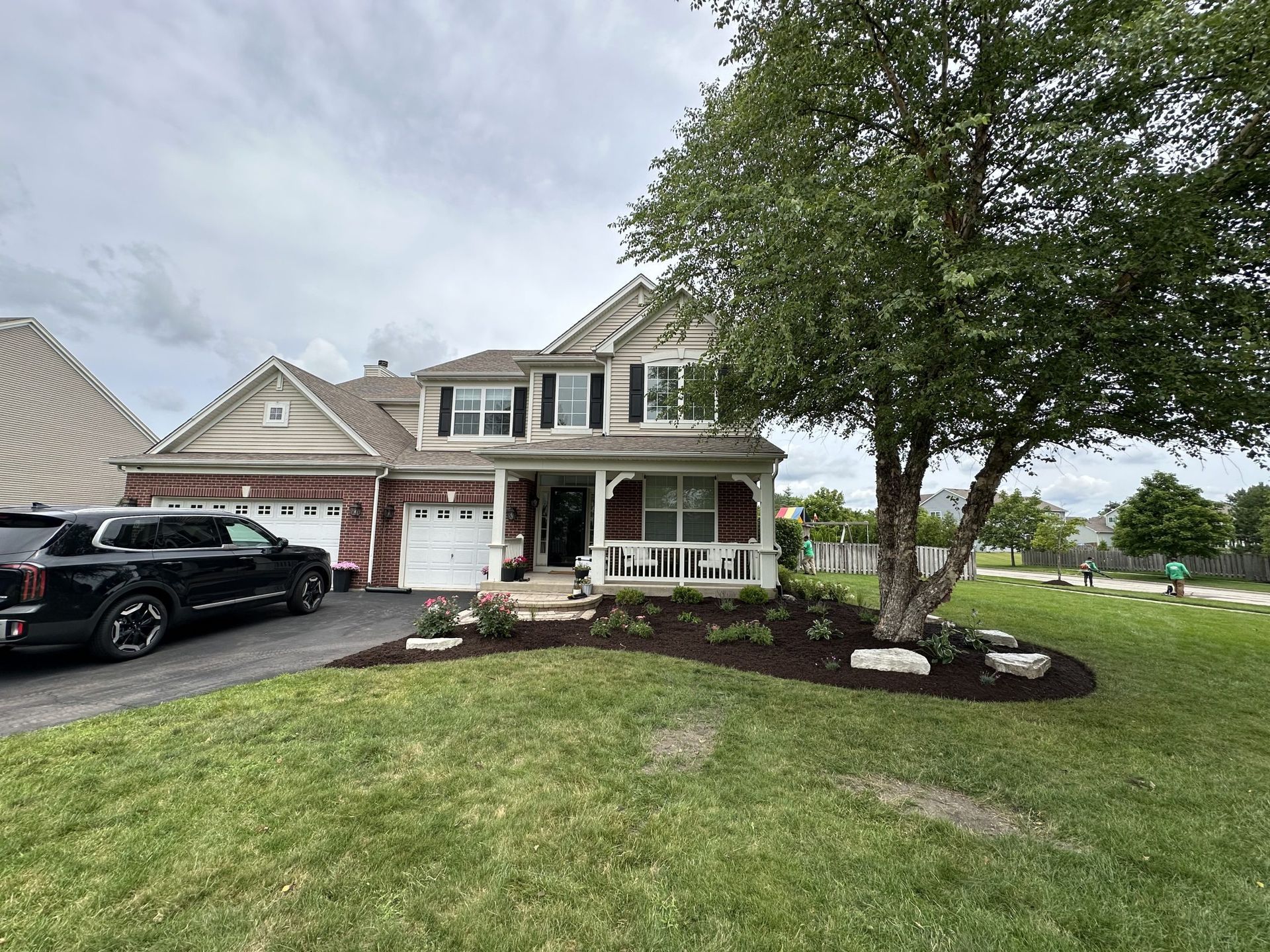 Two-story house with a black car parked in the driveway and a tree in the front yard.