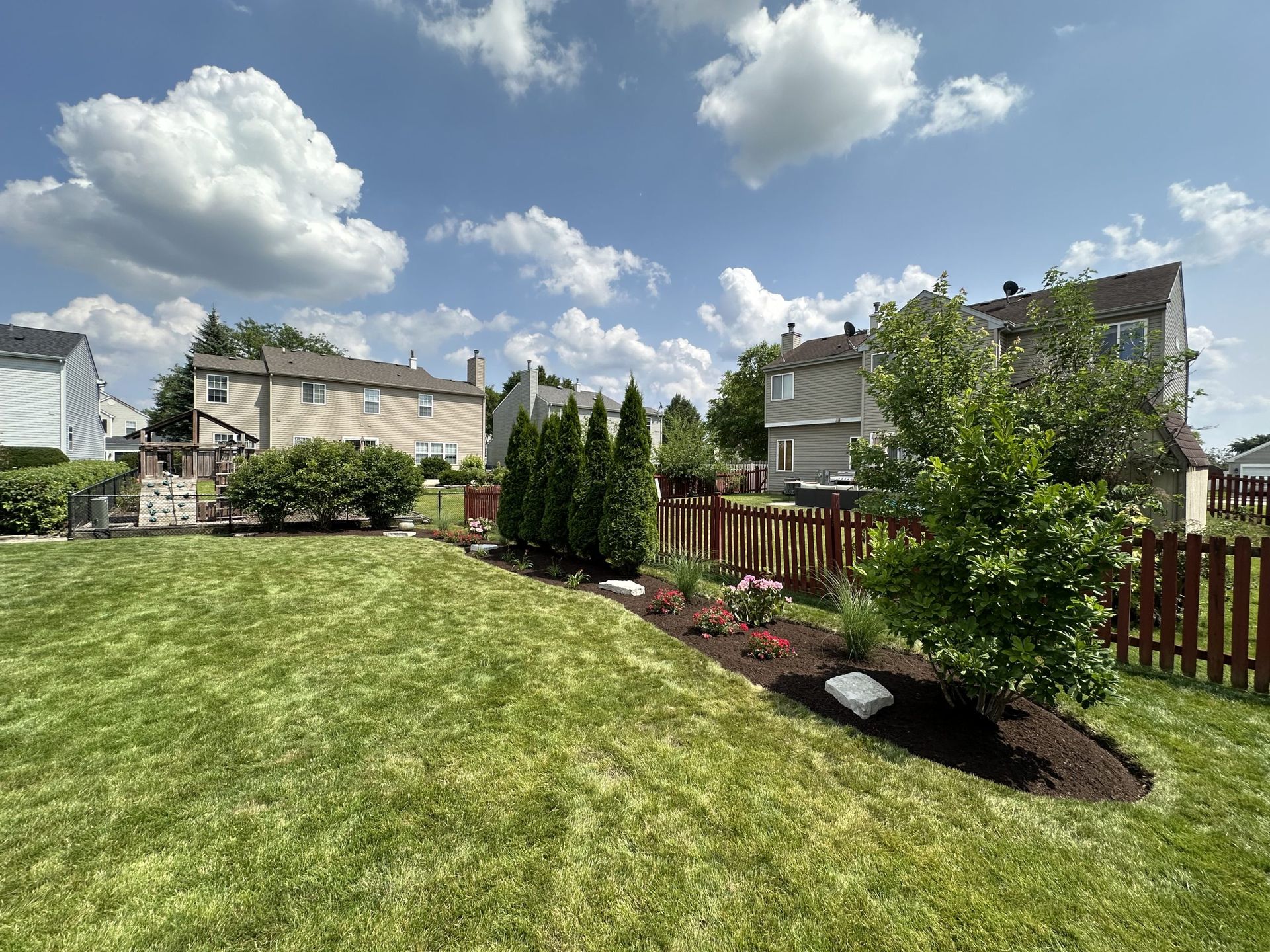 Lush backyard with green grass, brown mulch, red picket fence, and houses under a partly cloudy blue sky.