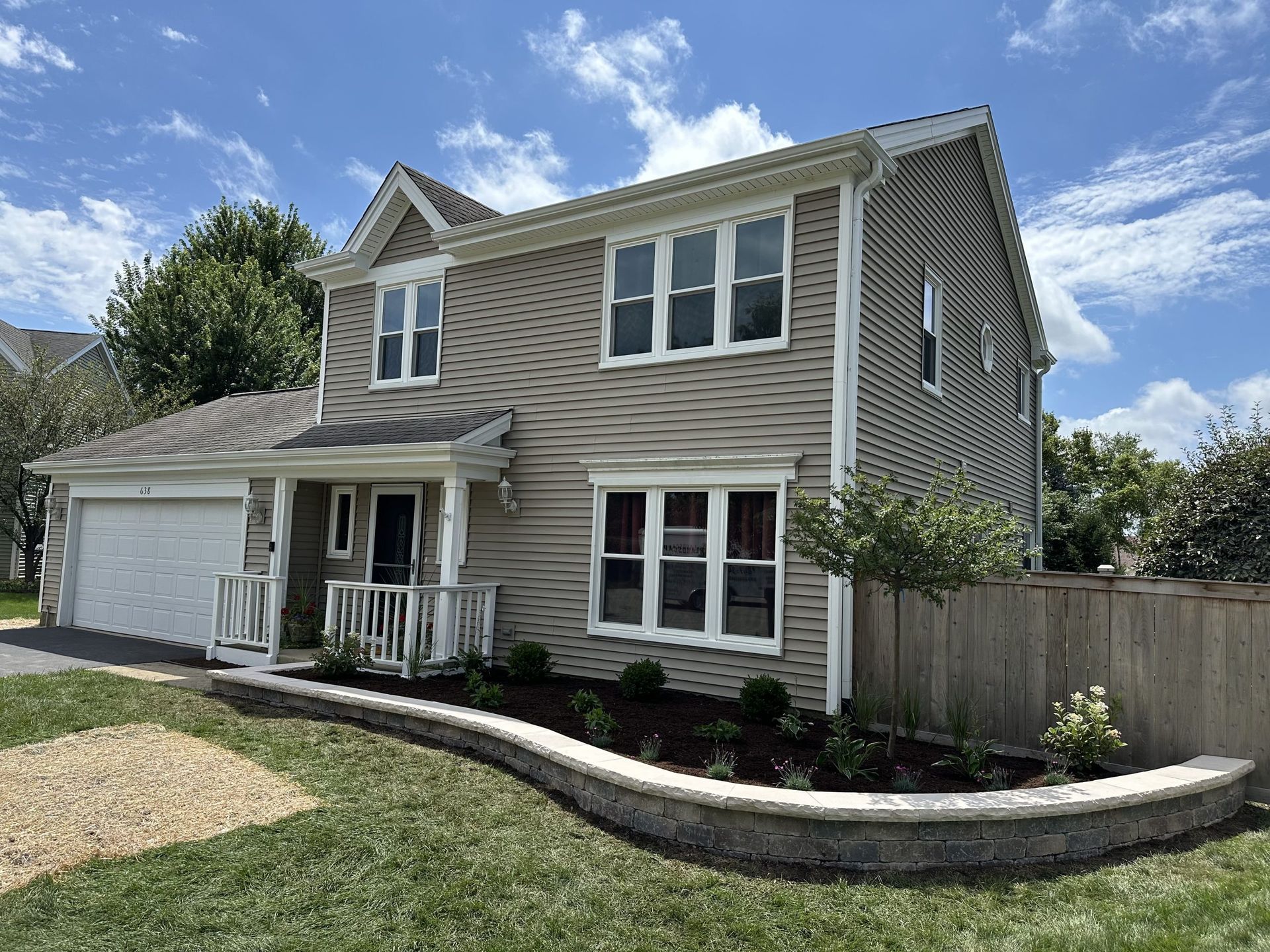 Two-story beige house with a garage, front porch, and a decorative landscaped retaining wall.