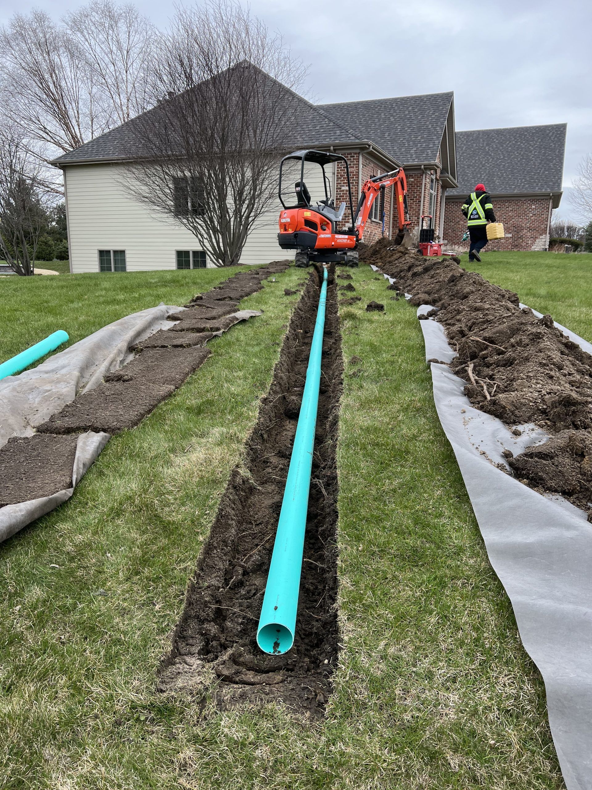 Trench in grass with blue pipe. Small excavator and worker near a house under construction.