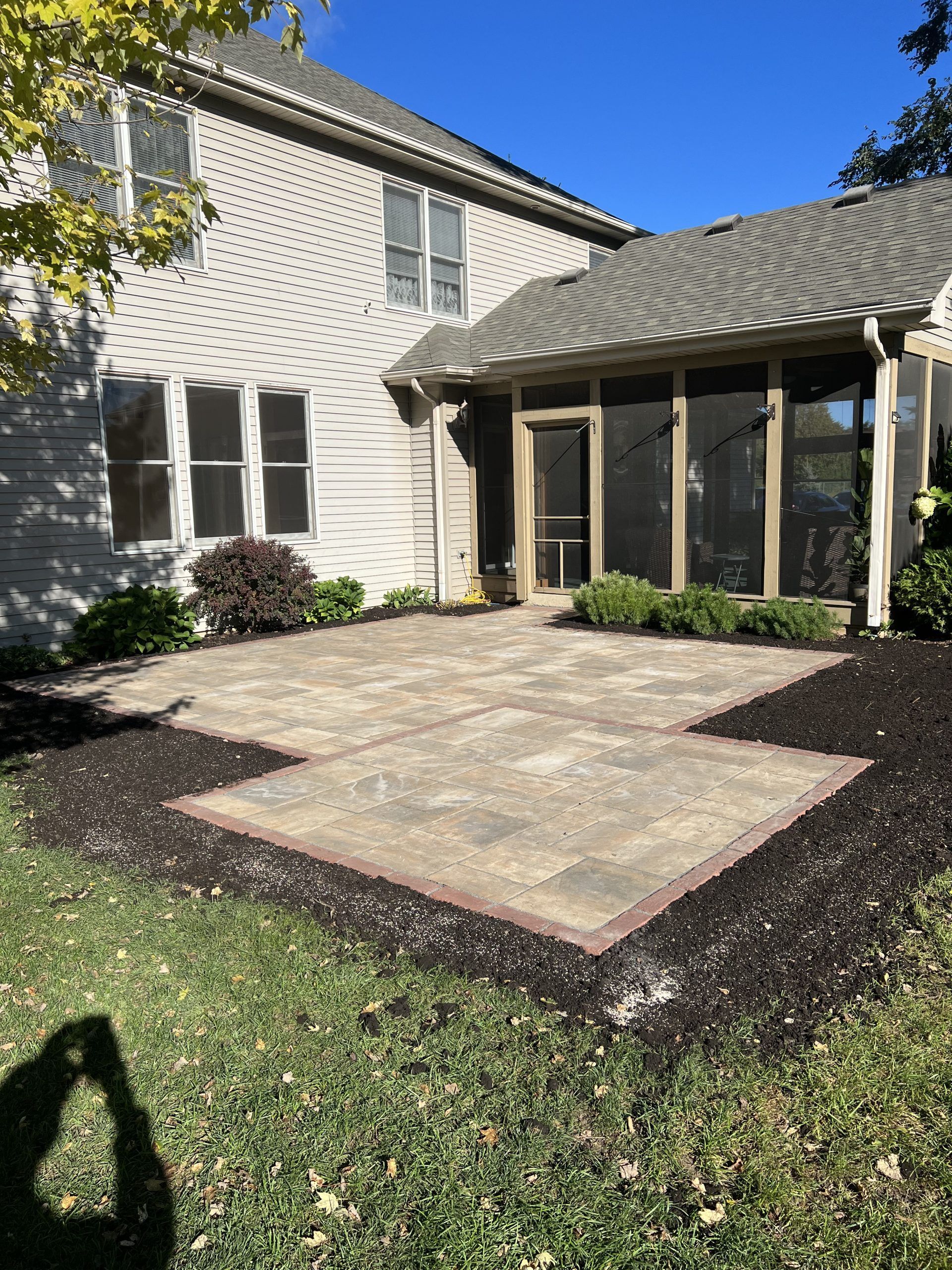 Backyard patio with pavers, surrounded by dark mulch, adjacent to a beige house with a screened porch.