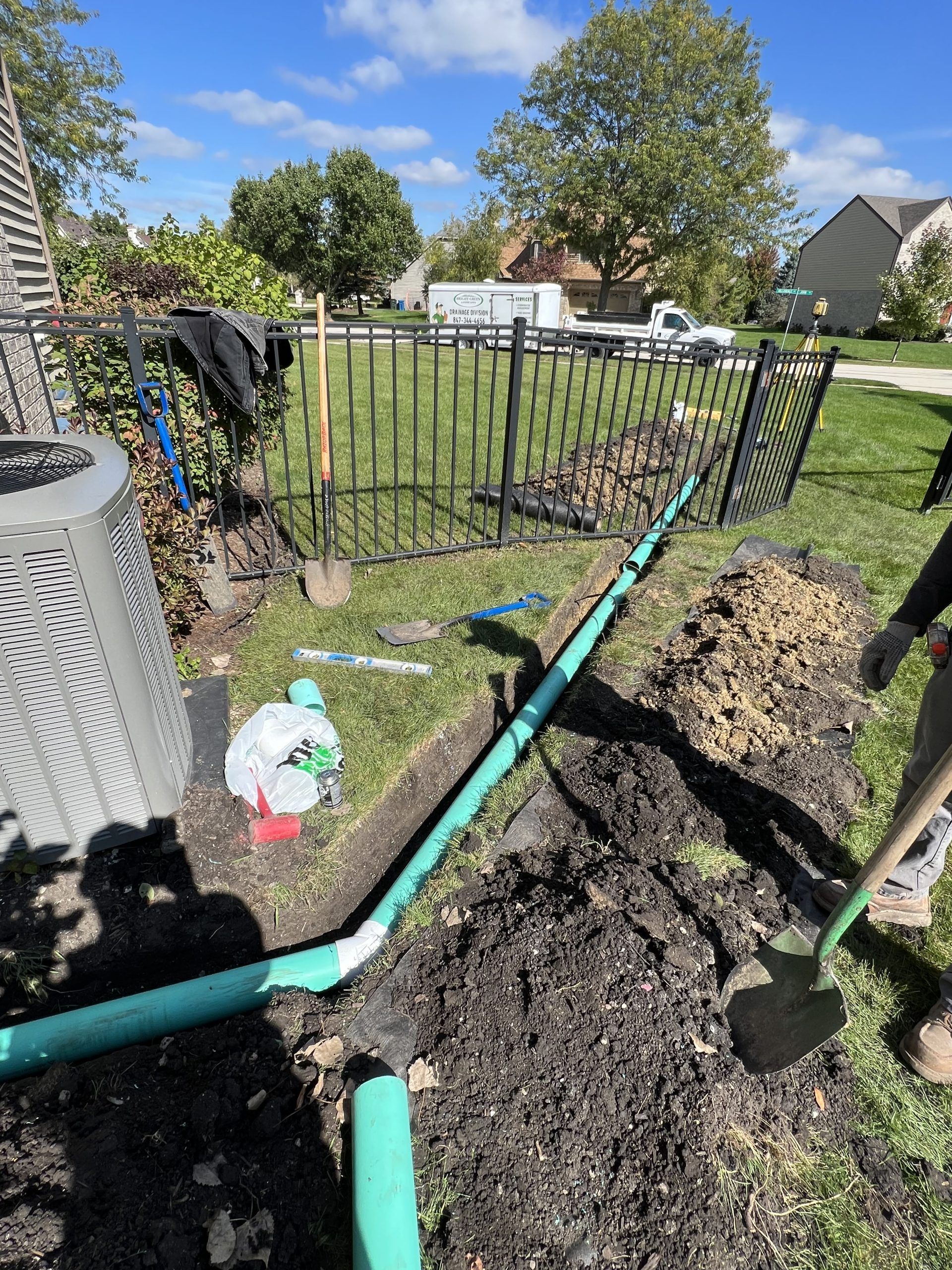 Green pipe being installed near a fence, shovels in dirt, sunny day.