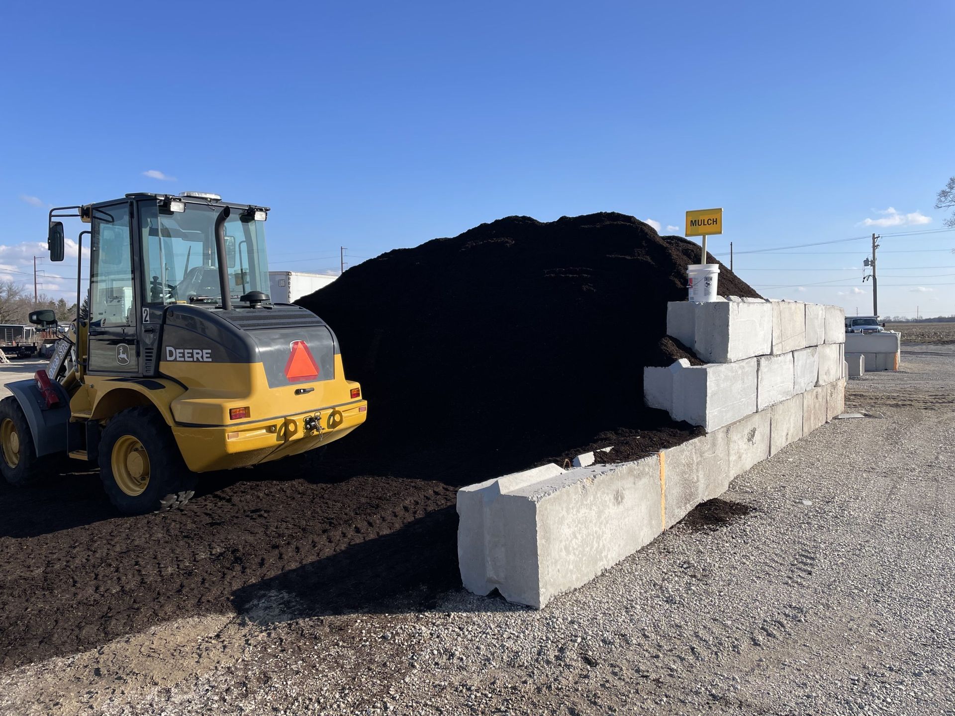 Yellow loader next to a pile of dark mulch, stacked concrete blocks, and a sign under a blue sky.
