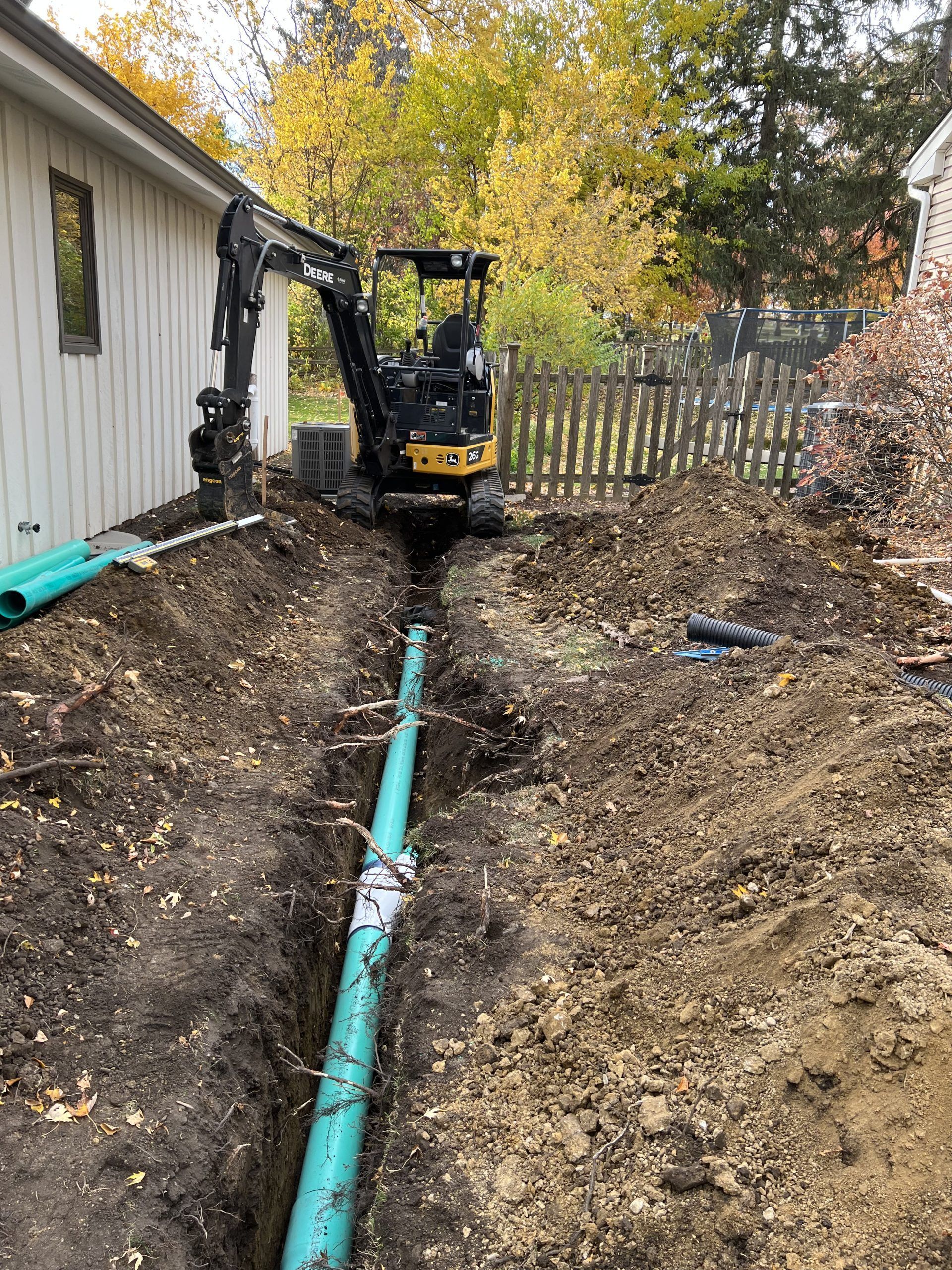 Mini excavator digging a trench beside a house, exposing a section of green pipe.