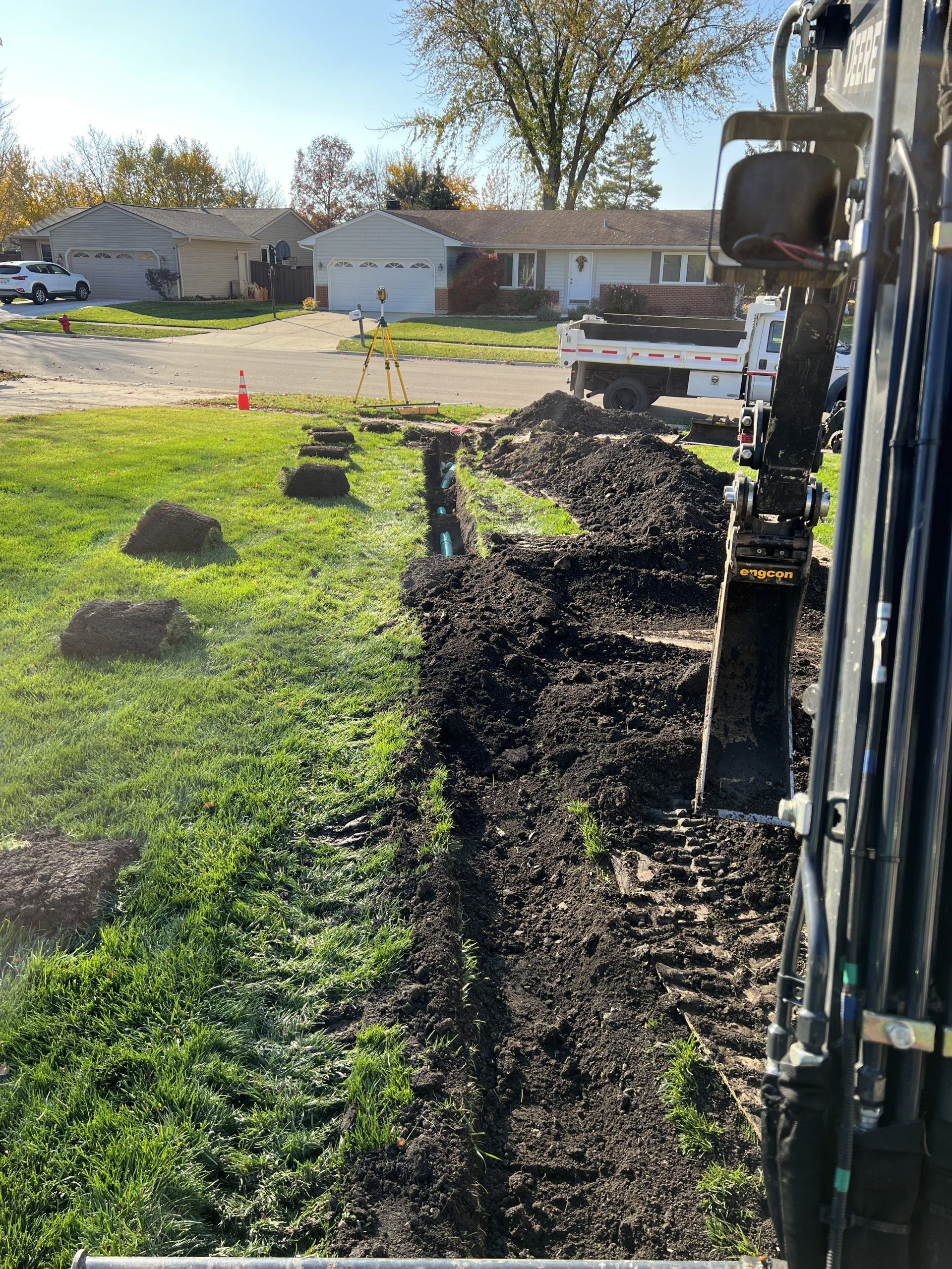 An excavator digging a trench along a grassy lawn in front of houses on a sunny day.