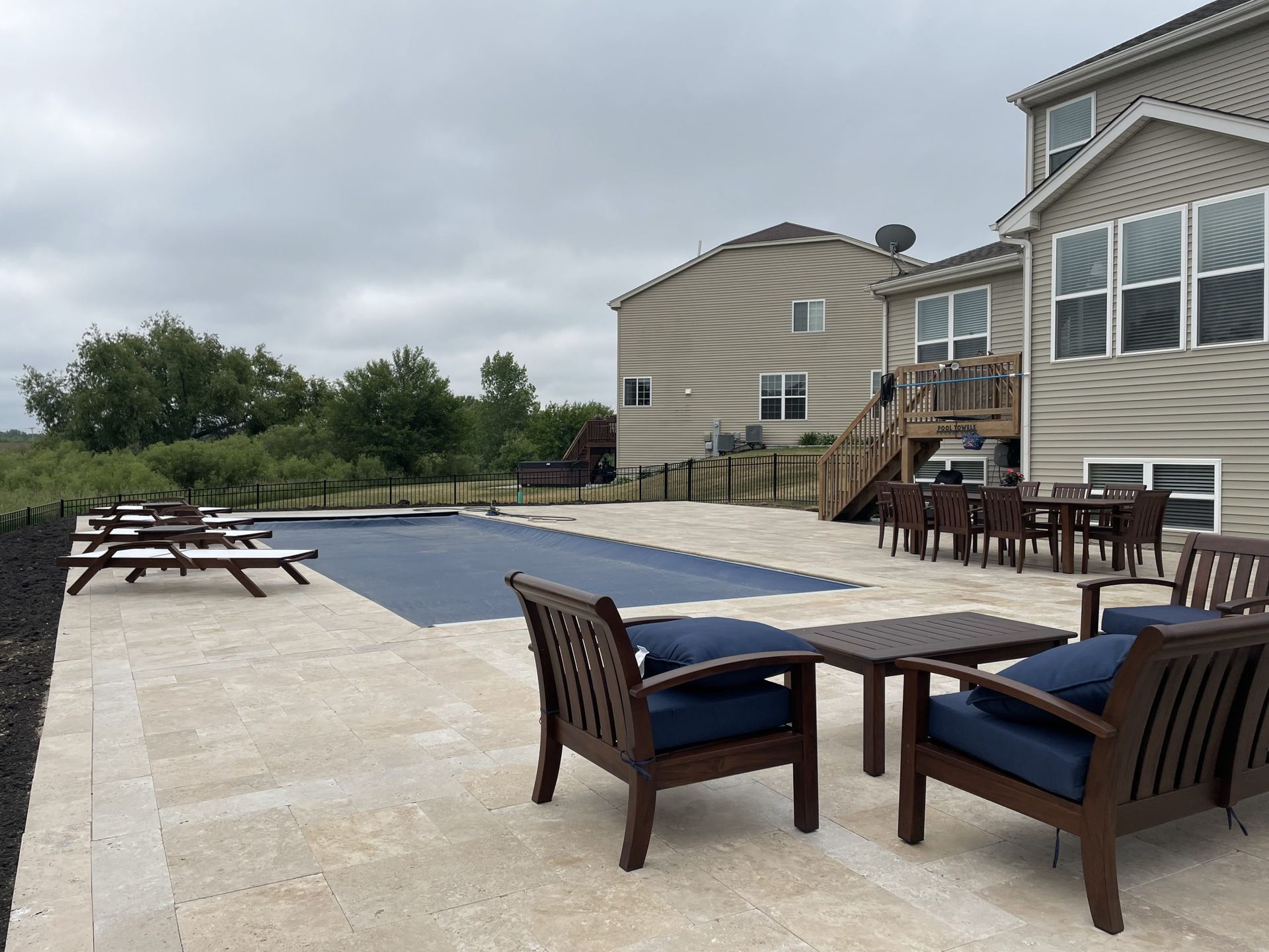 Backyard patio with pool, furniture, and house. Beige stone patio, blue cushions, cloudy sky.