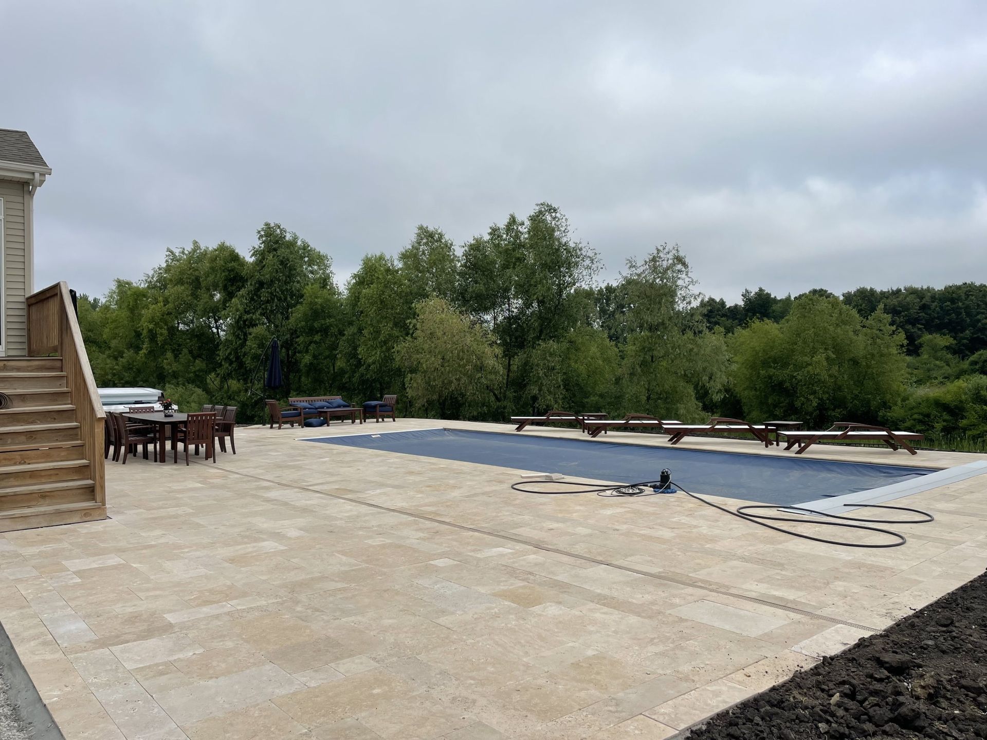 Backyard with rectangular pool covered by blue tarp, surrounded by beige patio, steps, and trees. Overcast sky.